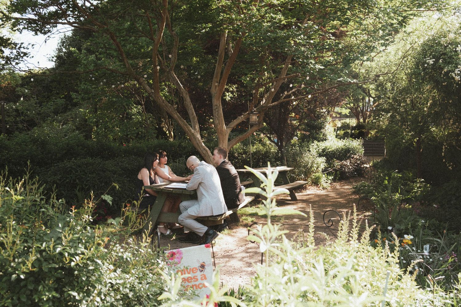 People sitting at a picnic table surrounded by lush green trees and plants in a serene garden setting.