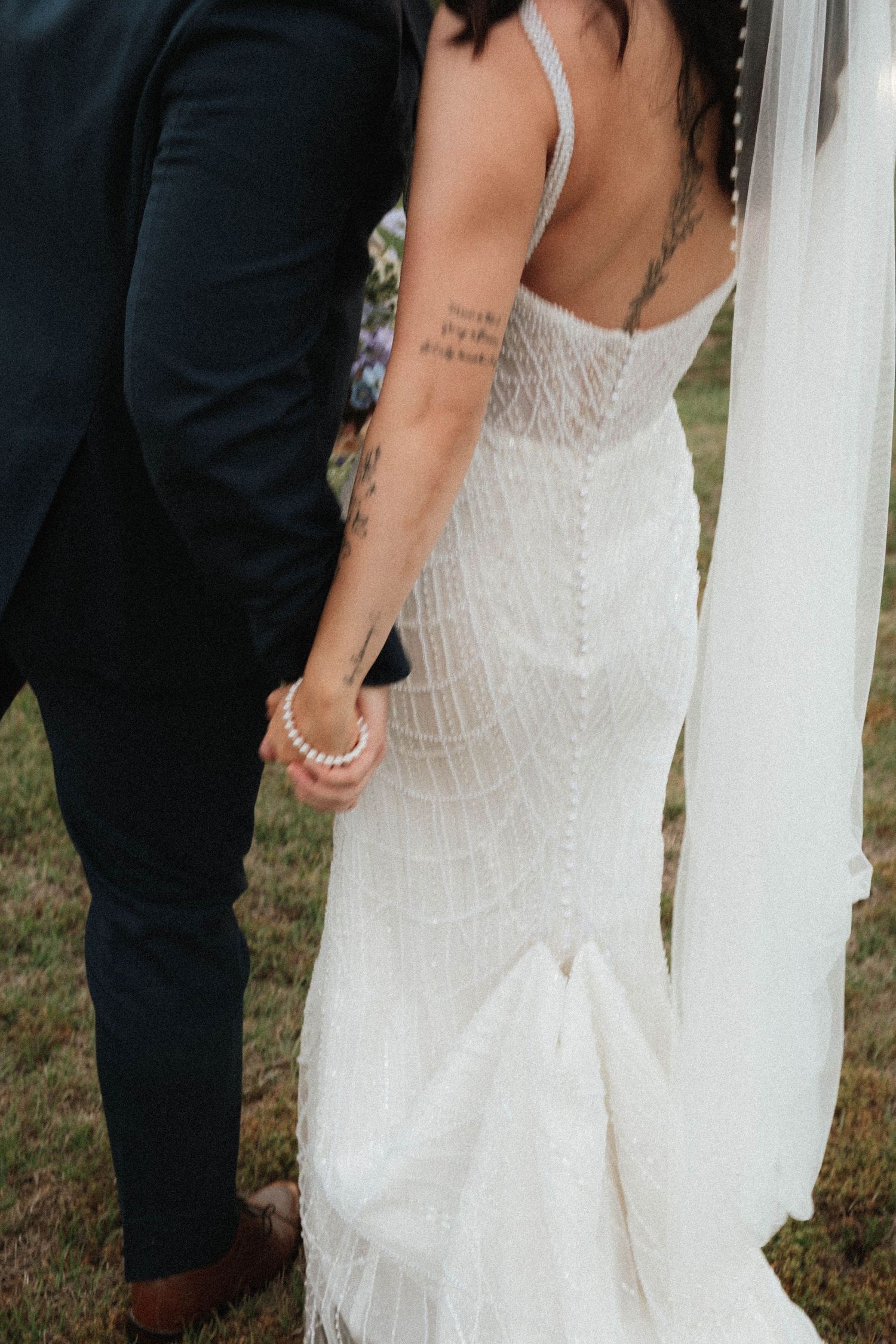 Bride in white lace mermaid wedding gown with veil, holding hands with groom in navy suit, back view.