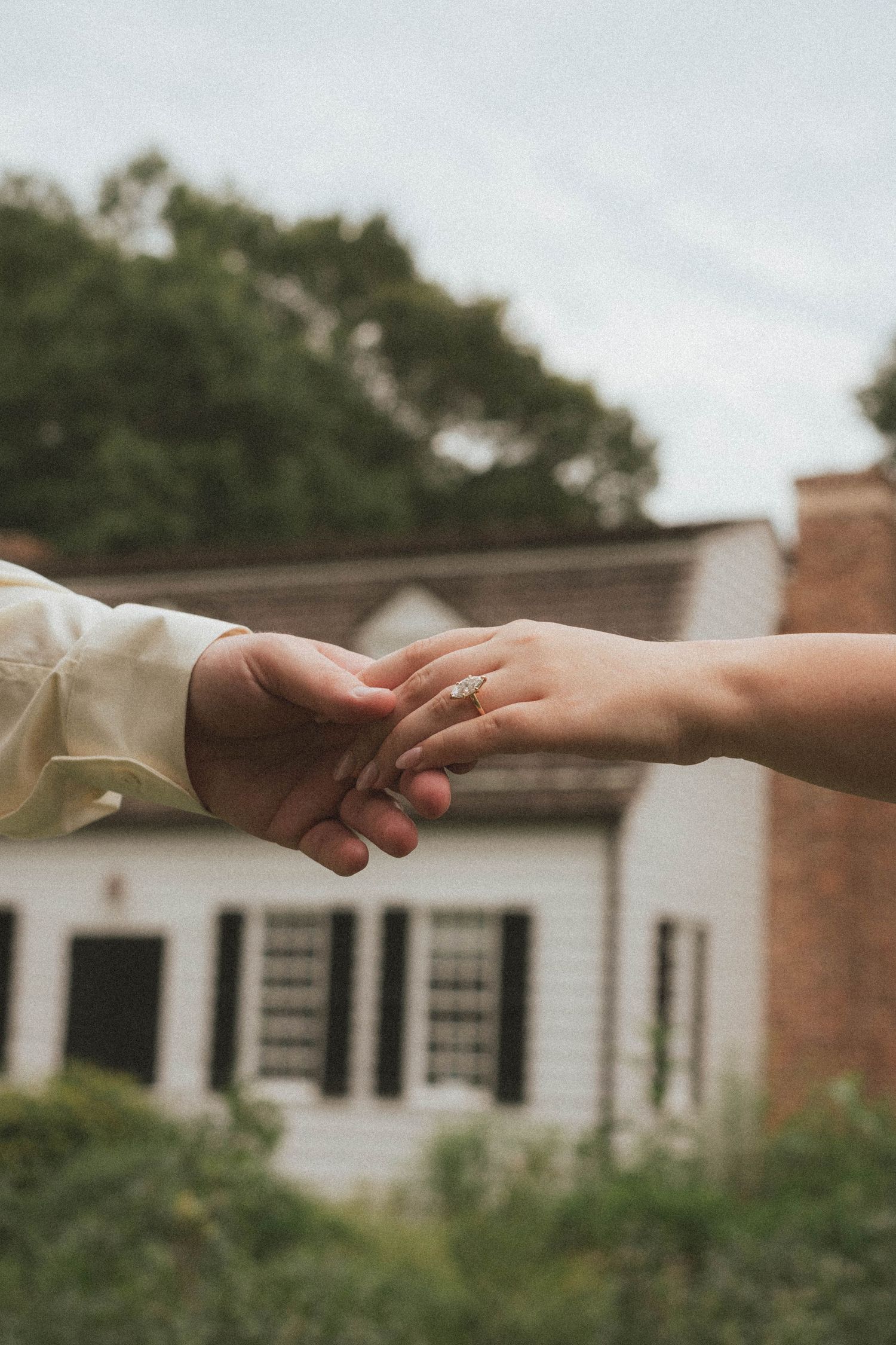 Couple holding hands outdoors with engagement ring visible, white colonial house and green trees in background.