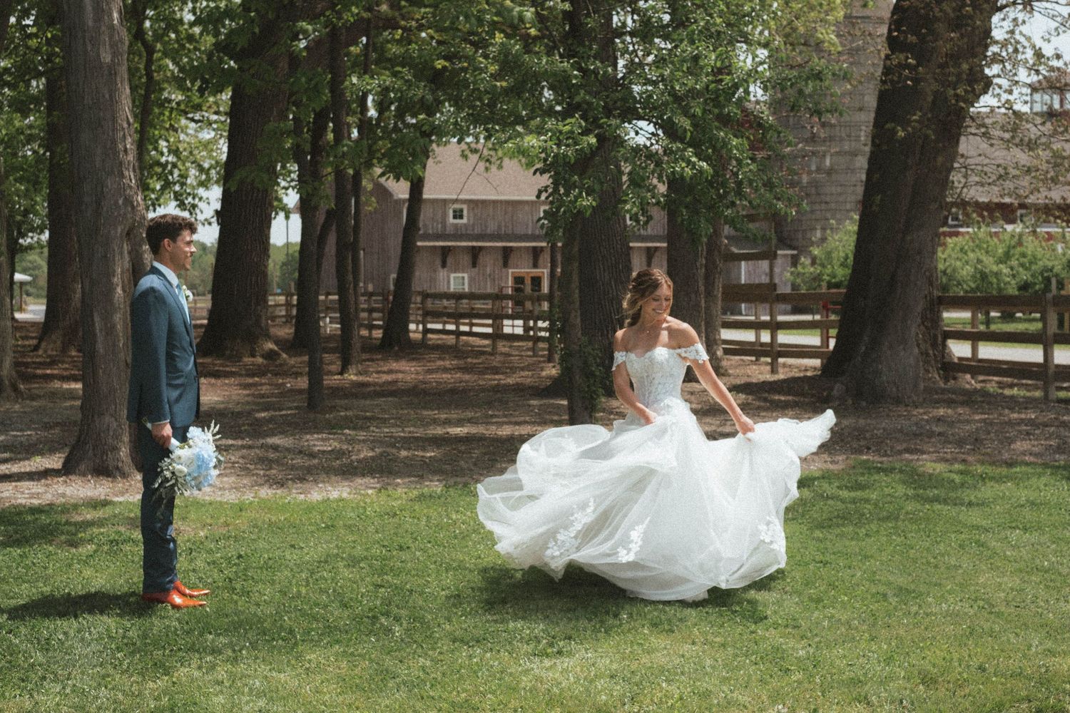 Bride in flowing white gown twirls for groom in blue suit during outdoor wedding photo session on lush green lawn.