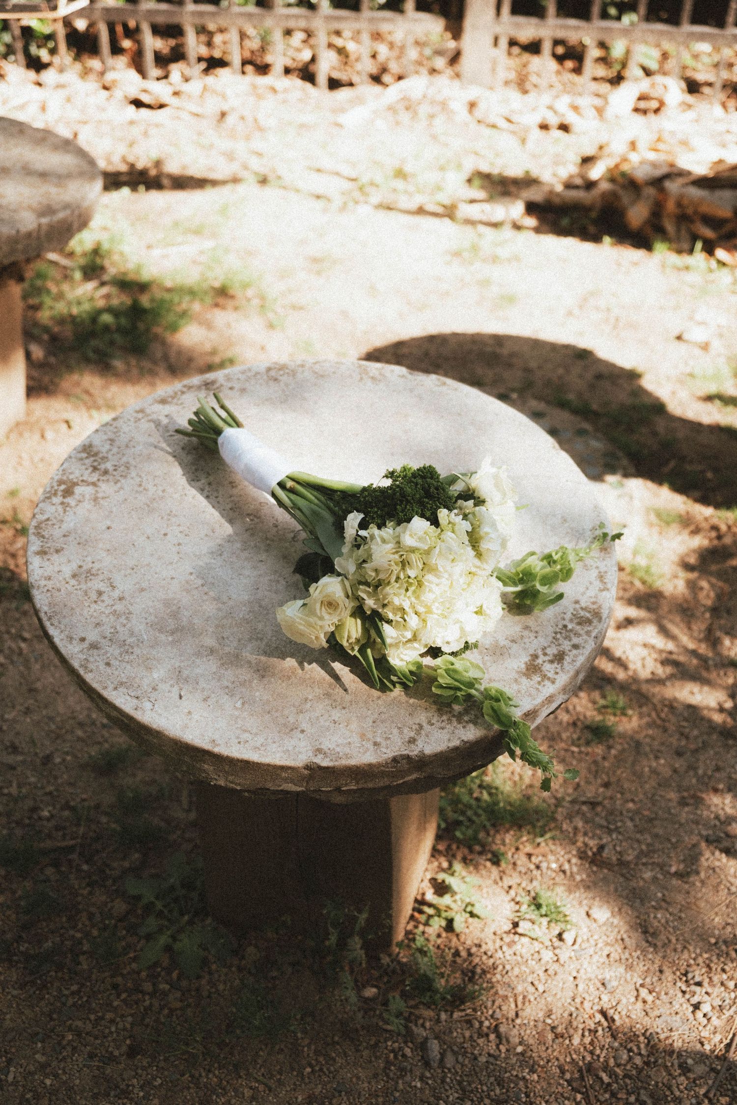 White wedding bouquet with roses and greenery resting on a rustic stone table outdoors in natural light.