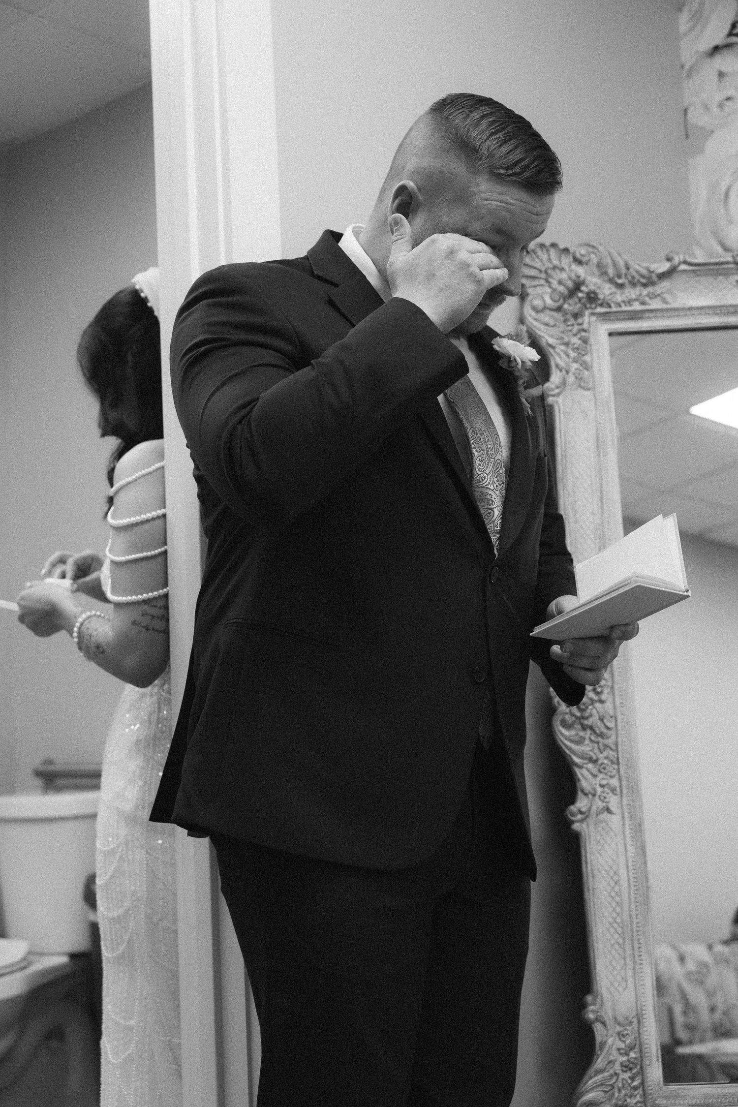 Emotional groom in black suit wiping tears while reading vows, bride visible in mirror reflection, black and white wedding photo.
