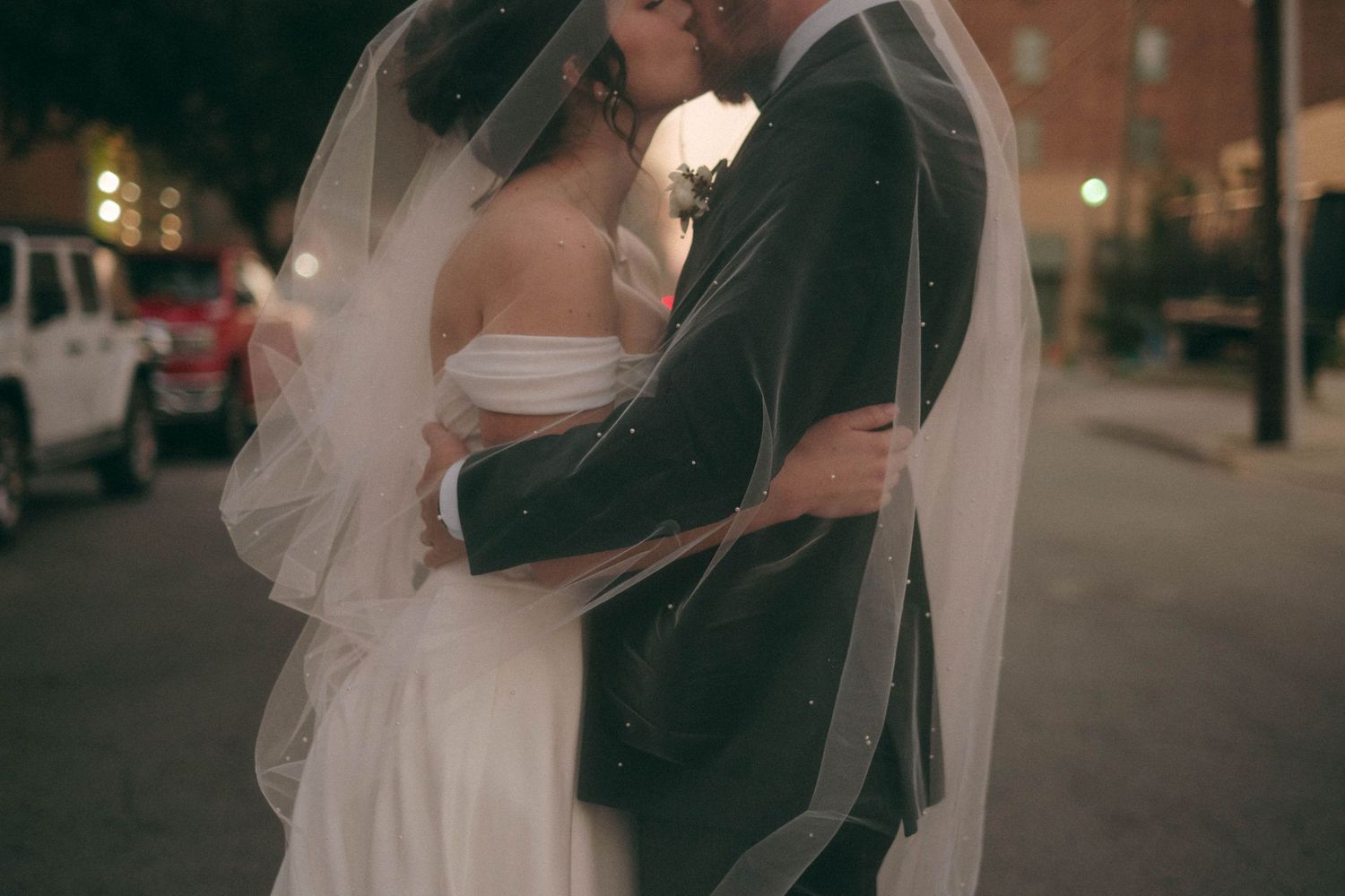 Wedding couple embracing under evening lights with bride's veil flowing.