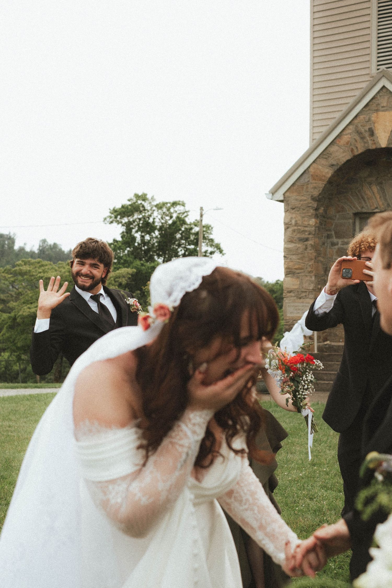 Emotional bride covering her face at outdoor wedding ceremony while groom waves happily near stone church.