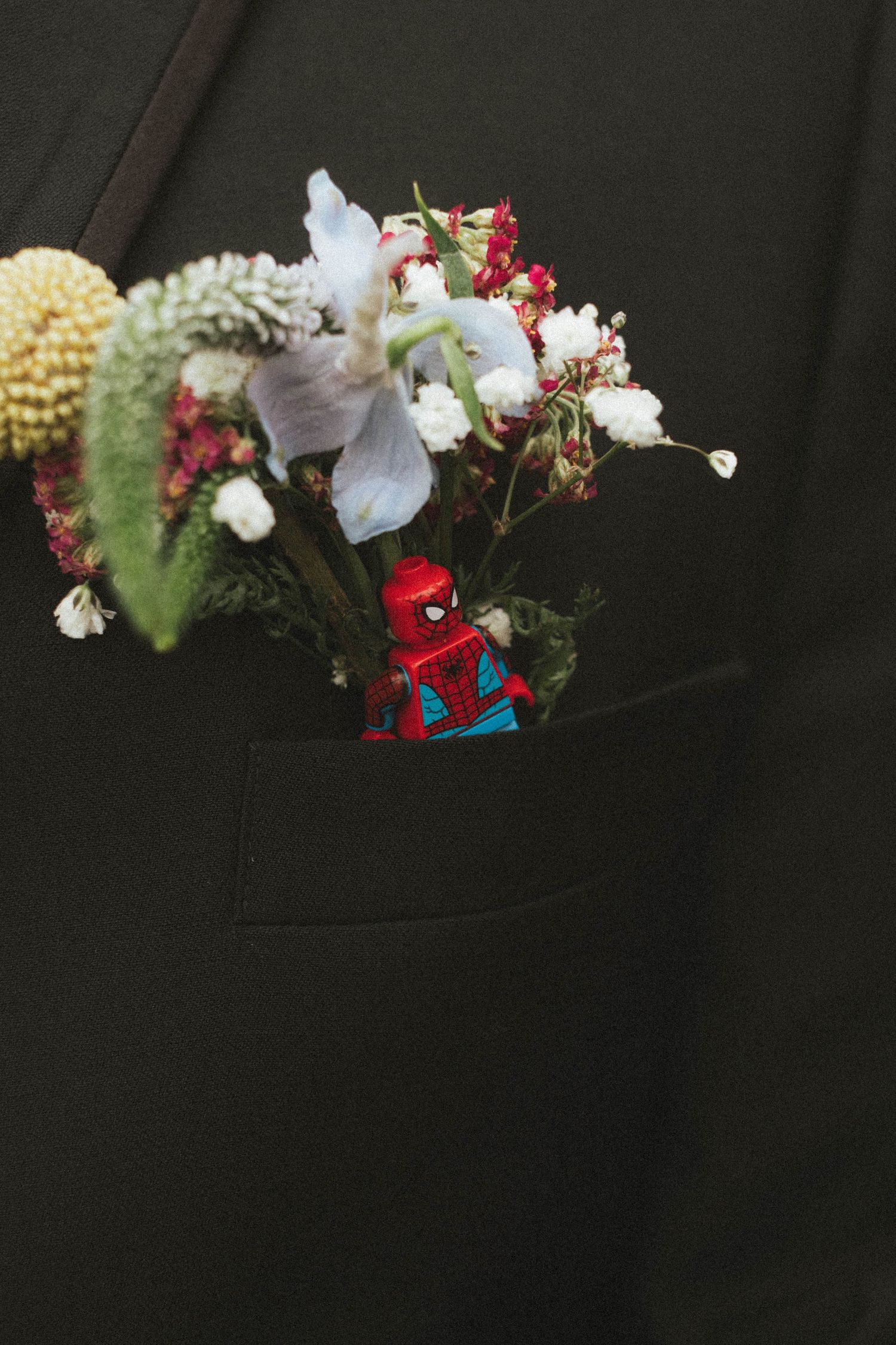 A floral boutonniere with a Spiderman LEGO figure tucked into a dark suit jacket pocket.