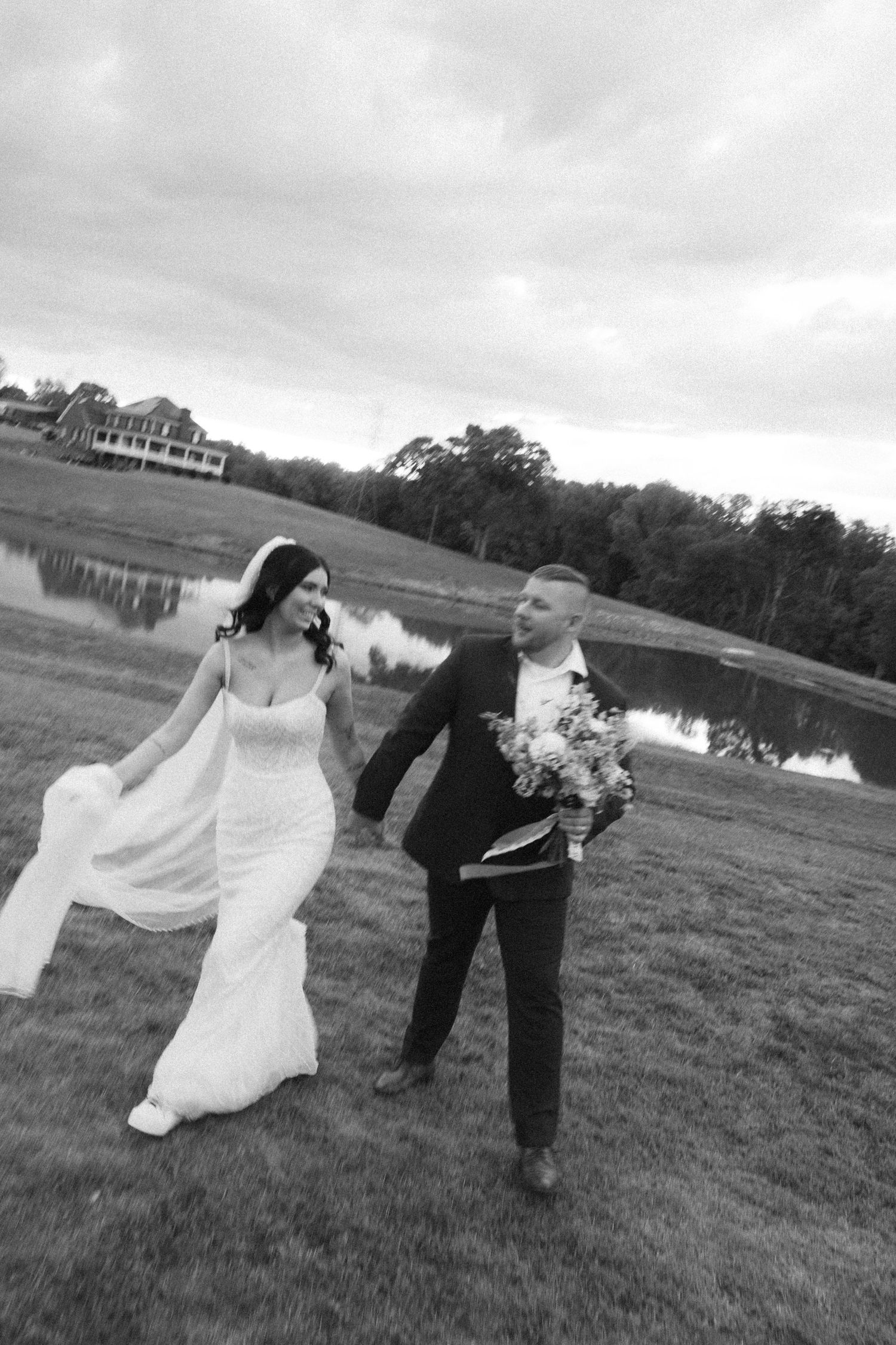 Black and white photo of a bride and groom walking hand in hand by a pond on their wedding day.