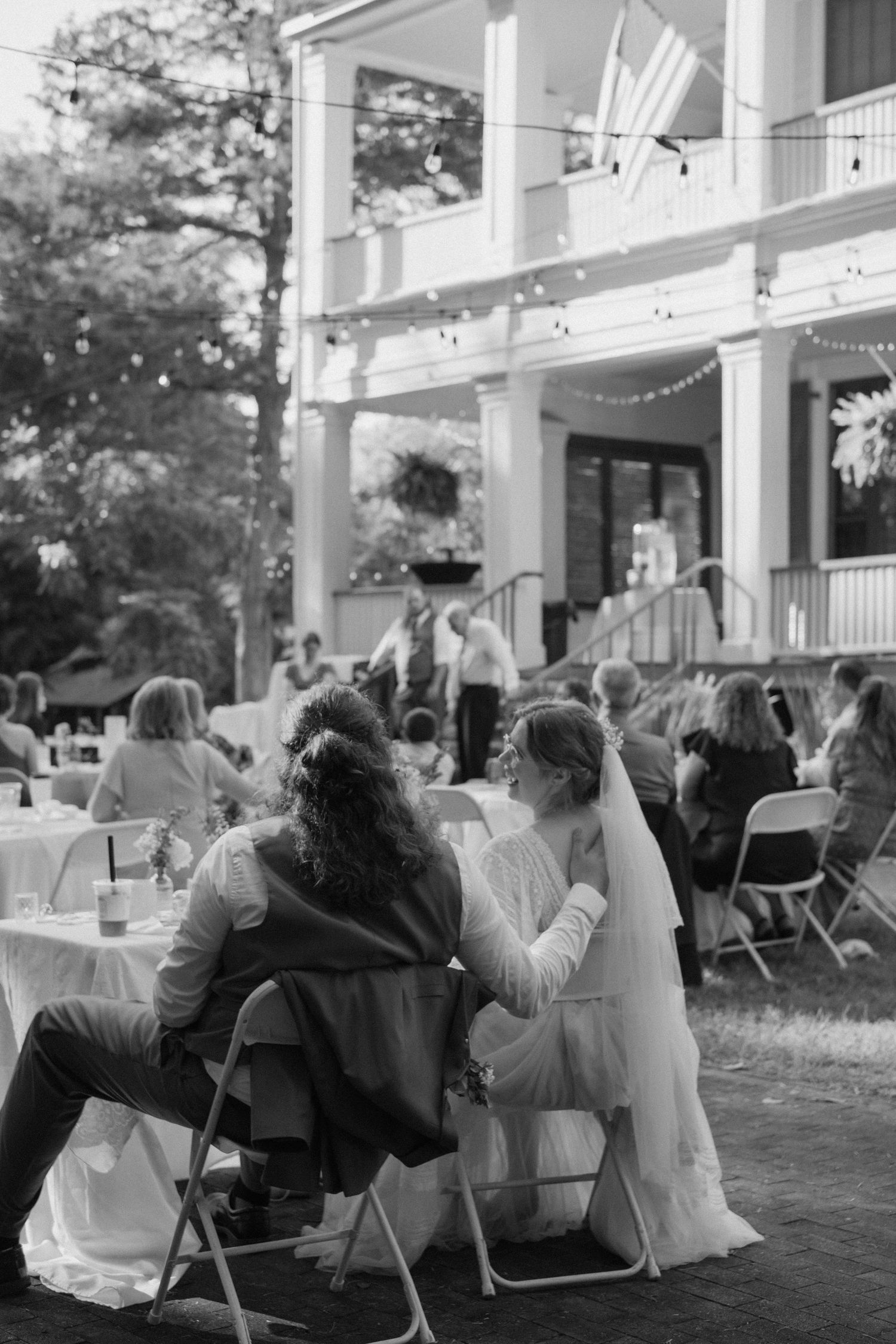 Wedding reception guests seated at tables on a porch with white chairs.