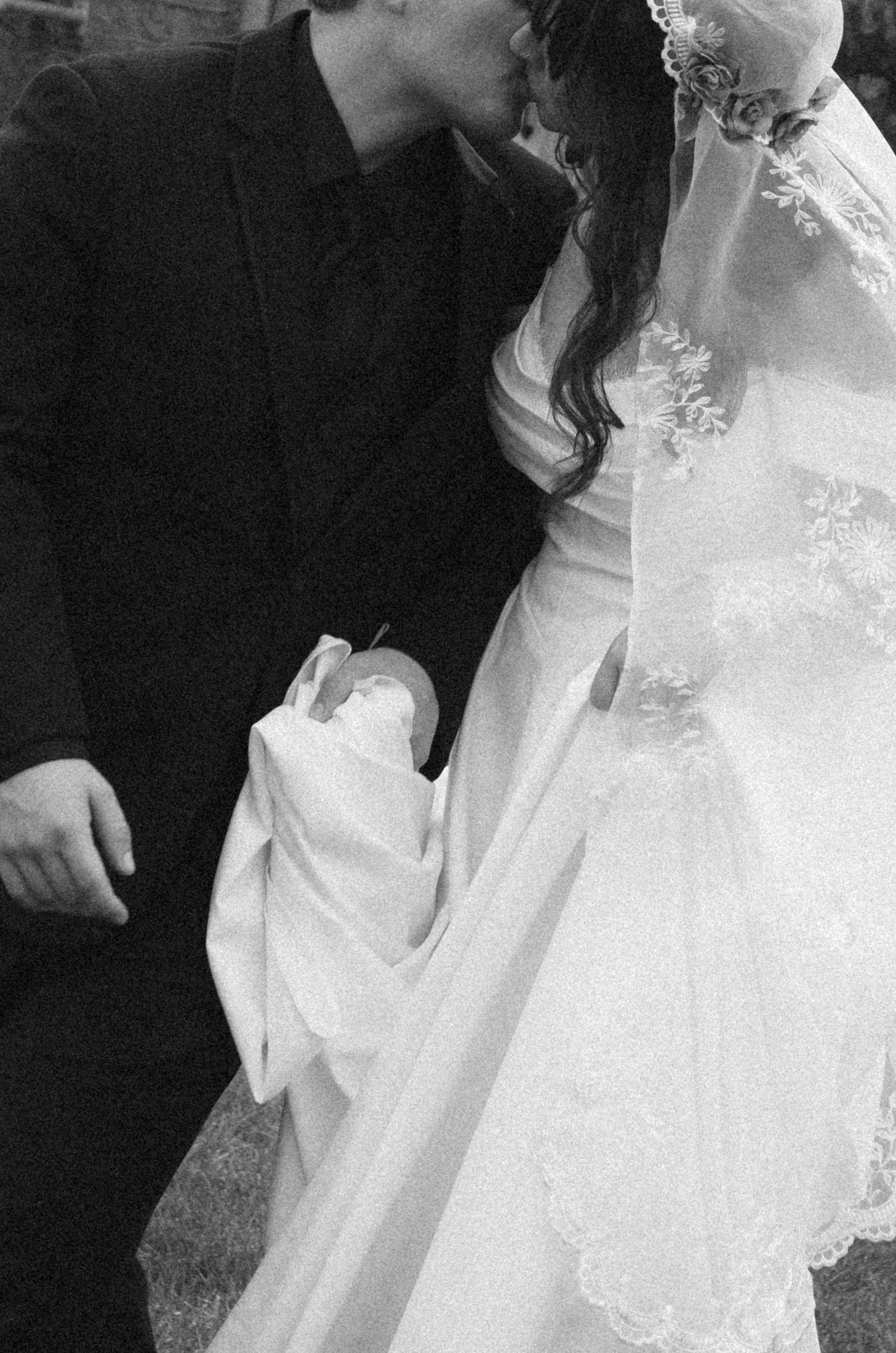 Black and white wedding photo of bride and groom kissing while holding a newborn baby in white blanket.