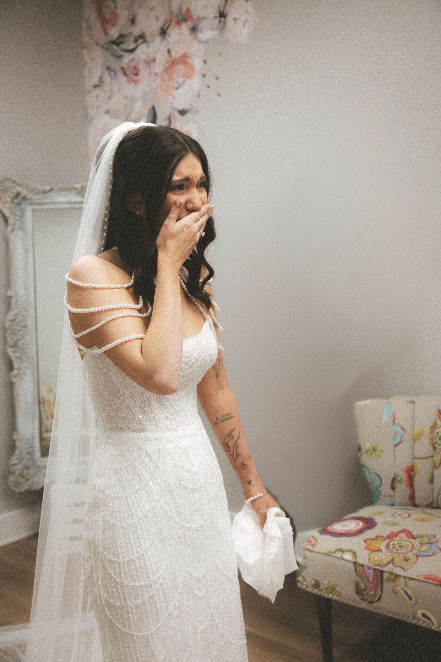 Emotional bride in white lace off-shoulder gown with veil, covering mouth in surprise, holding handkerchief.