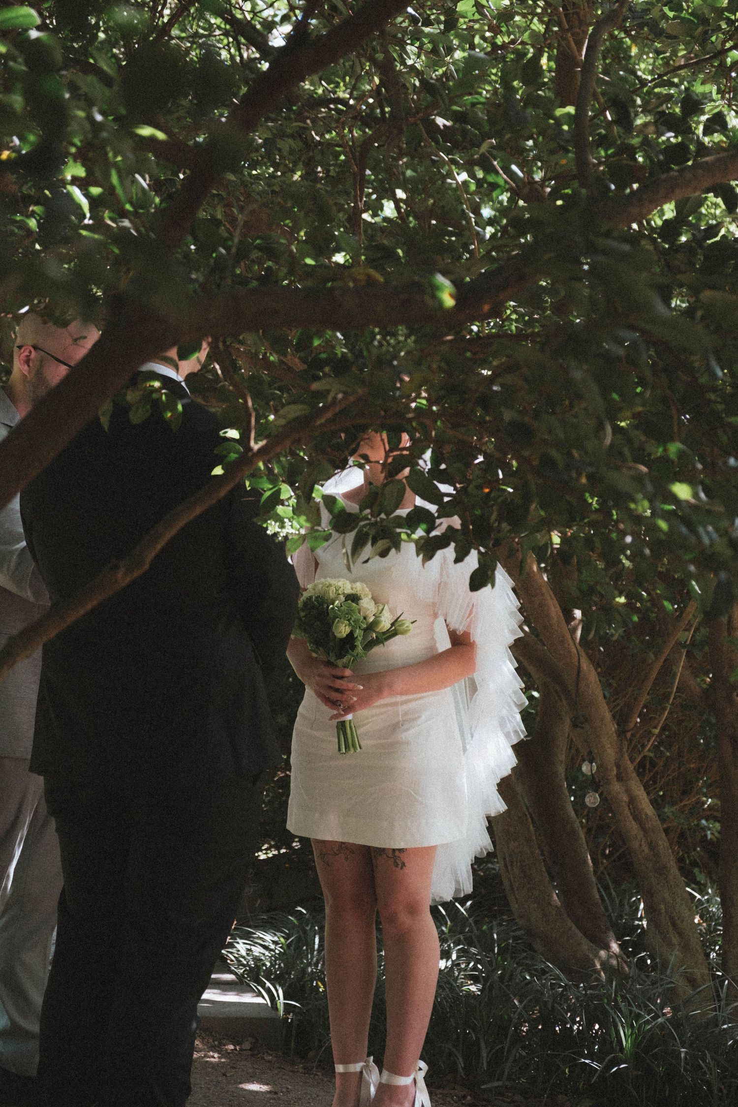 Bride in short white ruffled dress holding green bouquet stands under lush trees with groom during outdoor wedding ceremony.