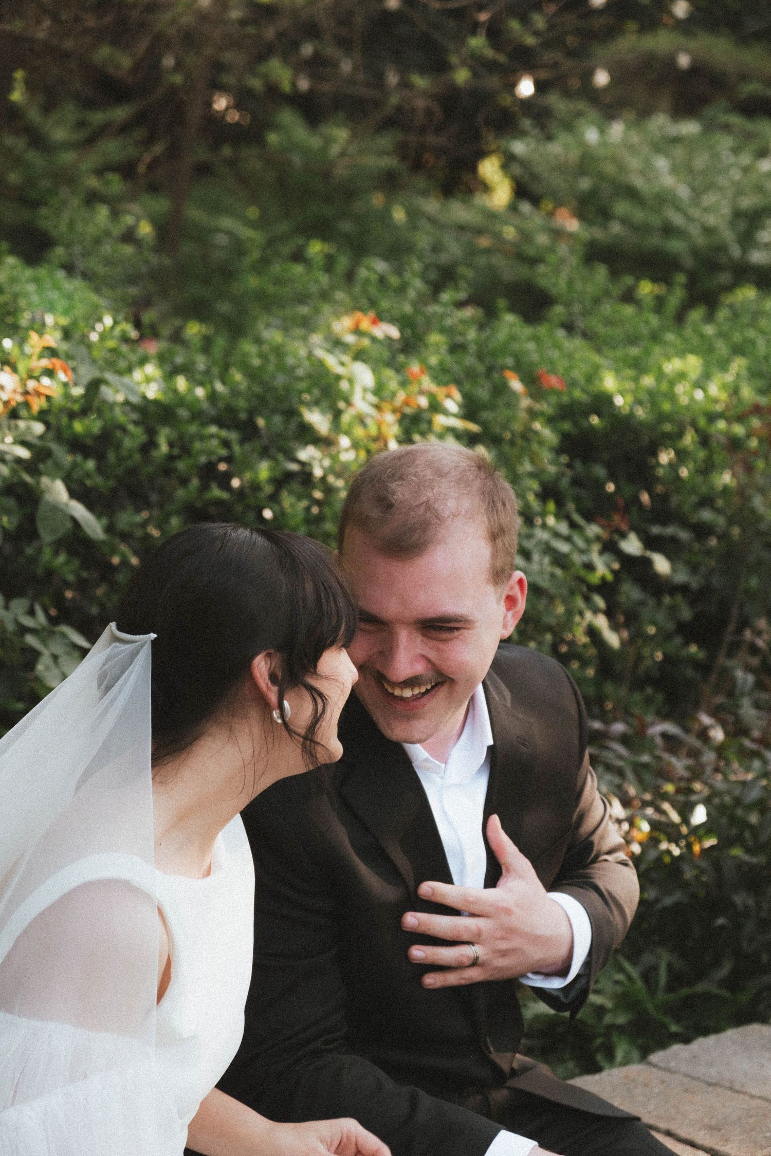 A joyful groom in a dark suit laughs with his bride in a white dress and veil during an outdoor wedding.