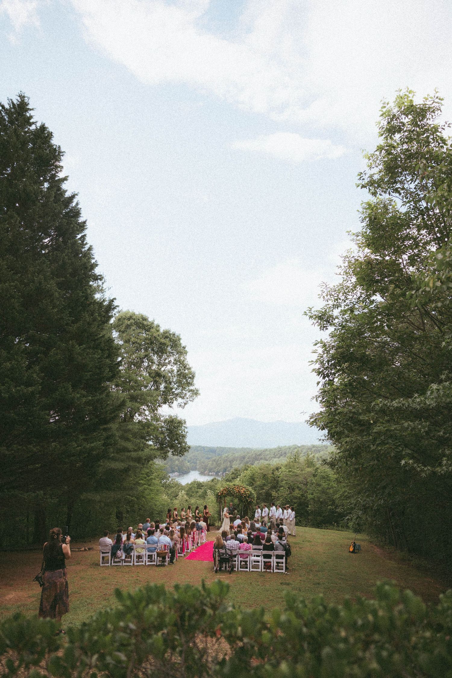 Outdoor wedding ceremony on a green hillside, framed by tall trees with mountain views and a pink aisle runner.