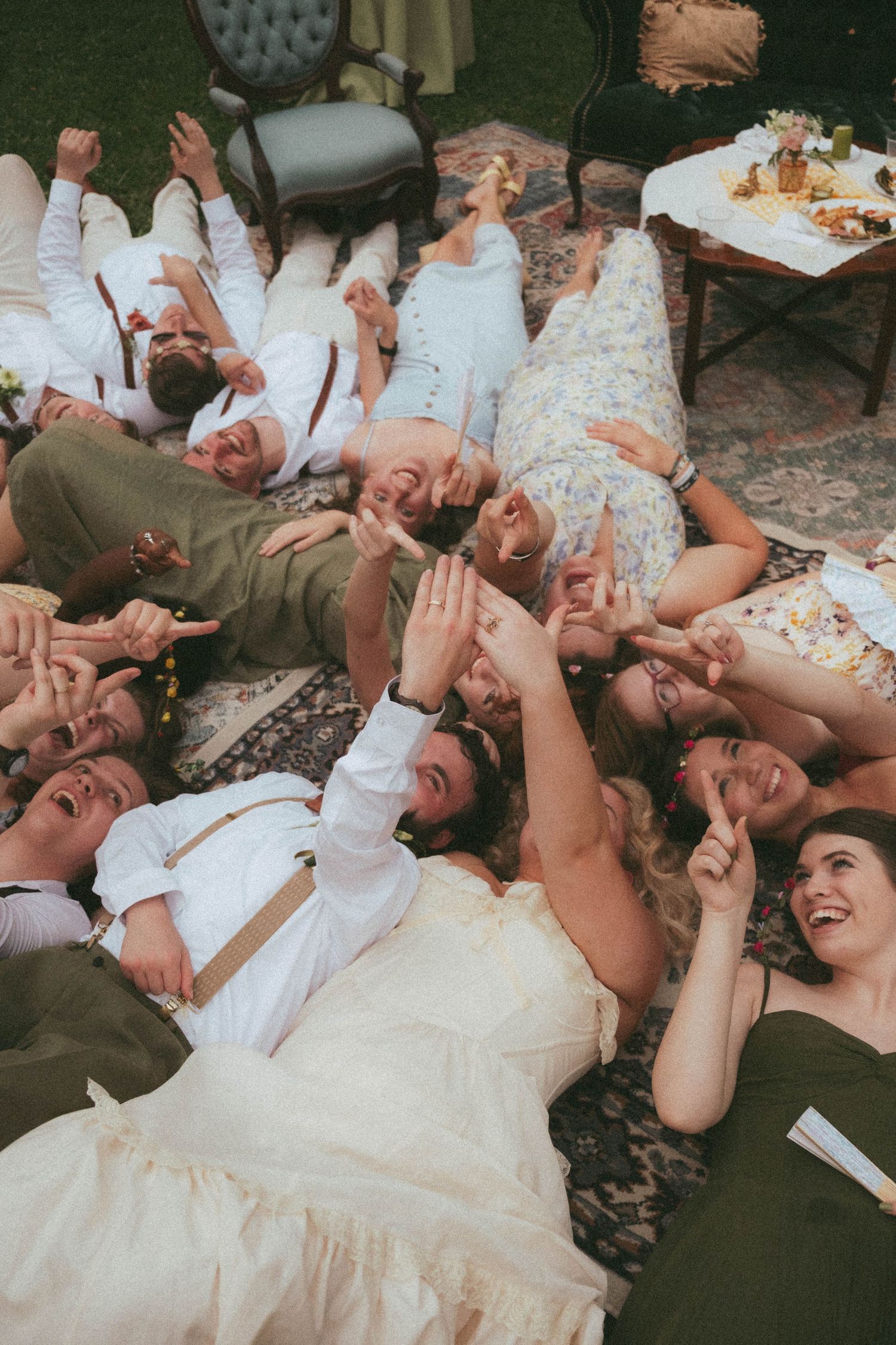Wedding group photo with bride and guests lying on floor laughing, pointing up, at outdoor reception.