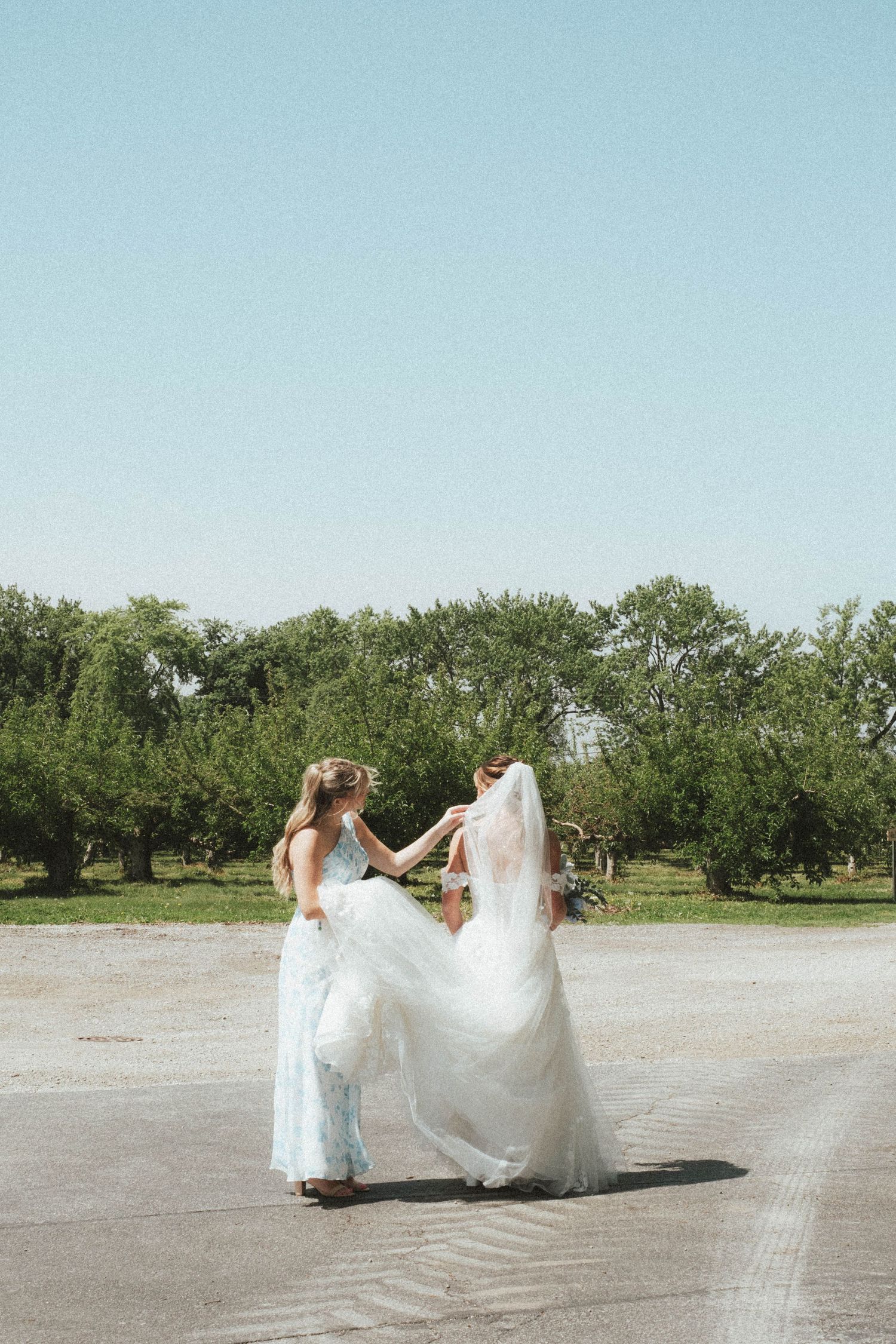 Bride in white gown dancing with bridesmaid in light blue dress outdoors on sunny day.