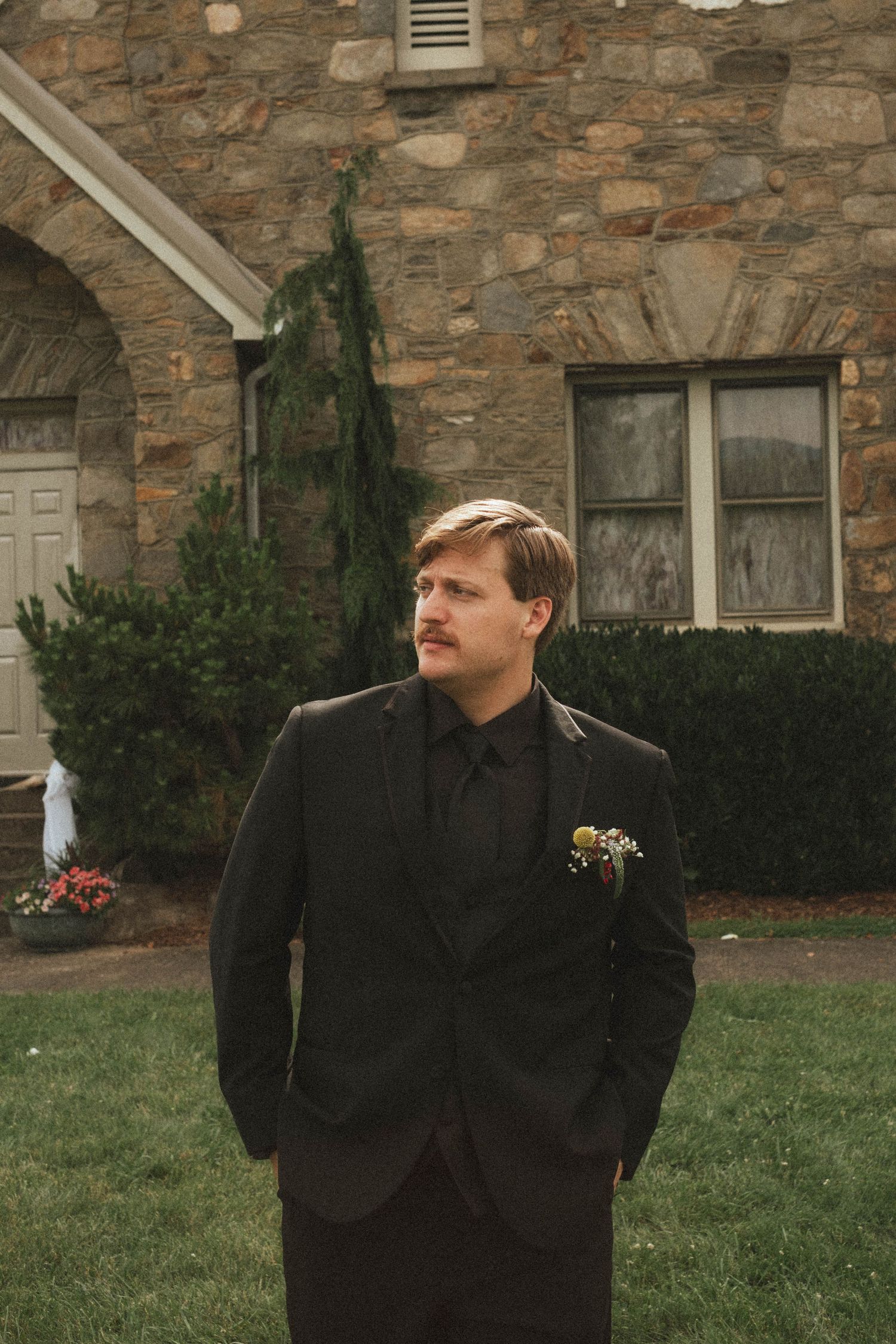 A groom in a dark suit with boutonniere stands on grass before a stone building.