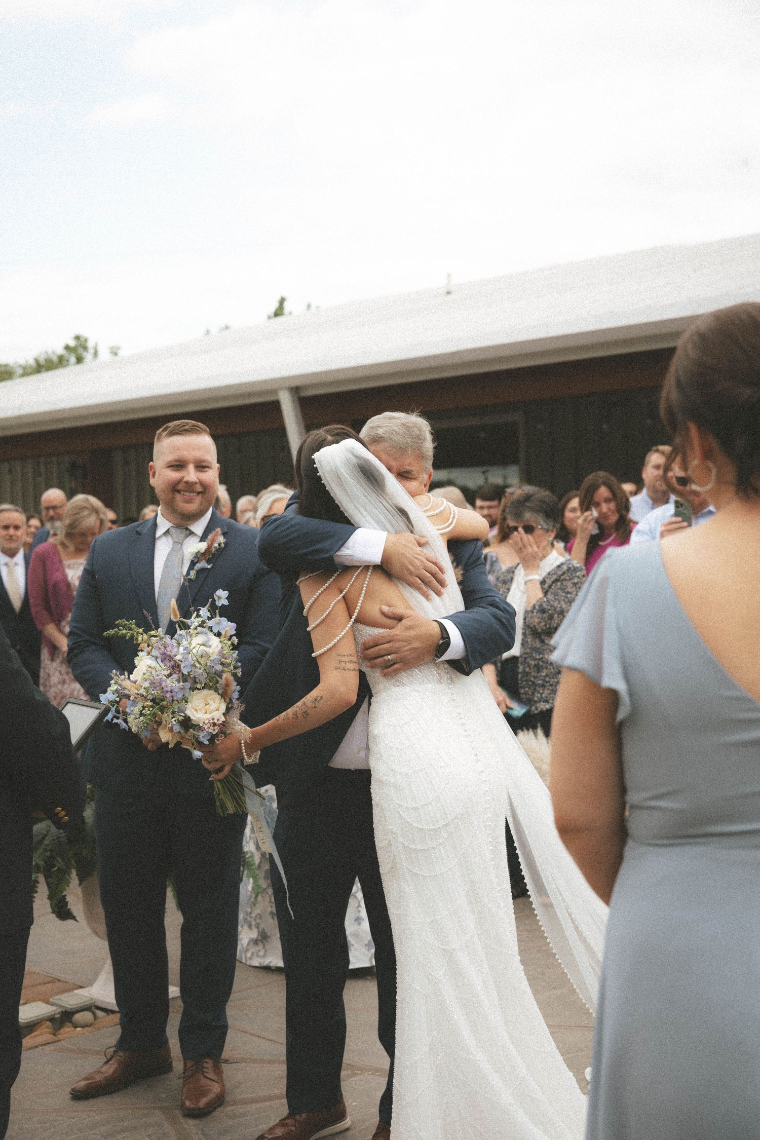Bride in lace gown hugging father after wedding ceremony while groom smiles holding bouquet outdoors.