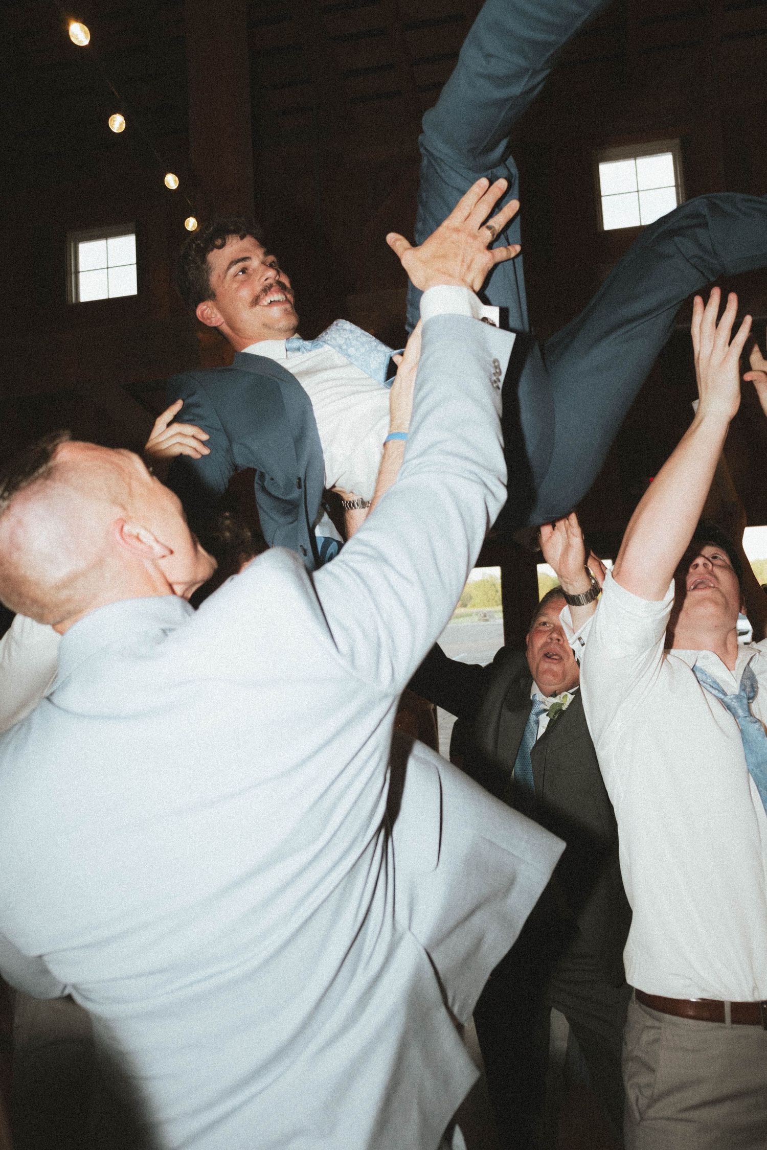 Groomsmen joyfully tossing the groom into the air during a lively wedding reception celebration.