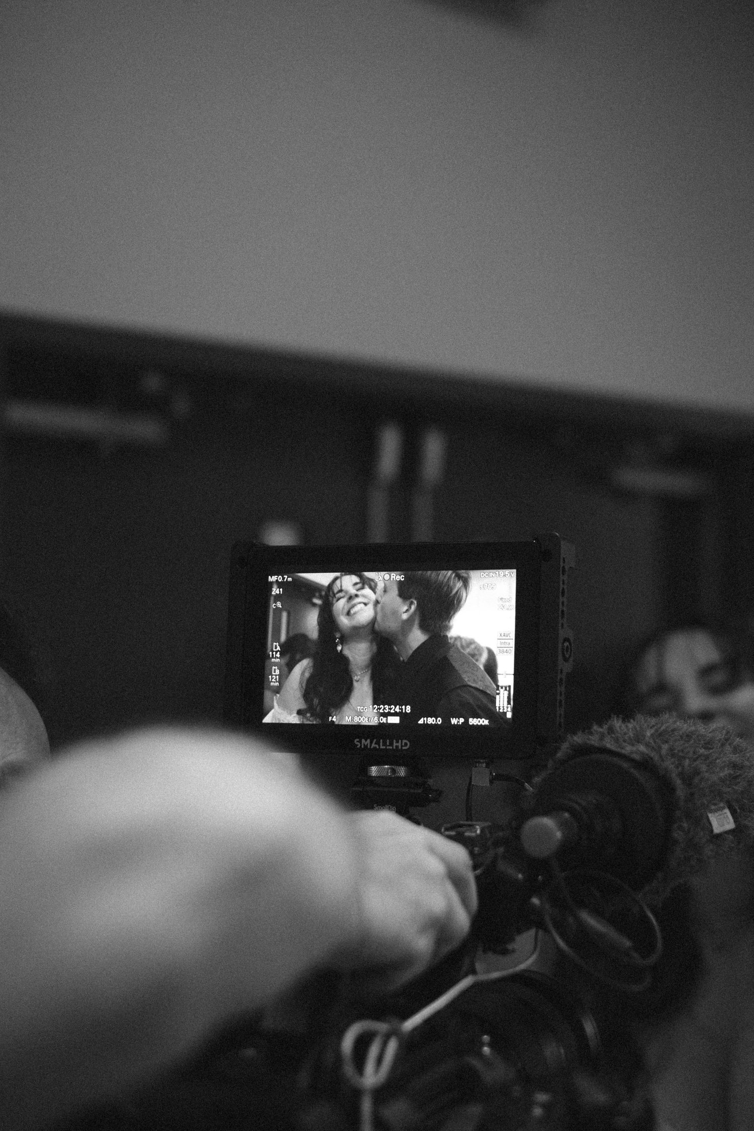 Black and white behind-the-scenes film set photo showing camera monitor displaying two people sharing an intimate moment.