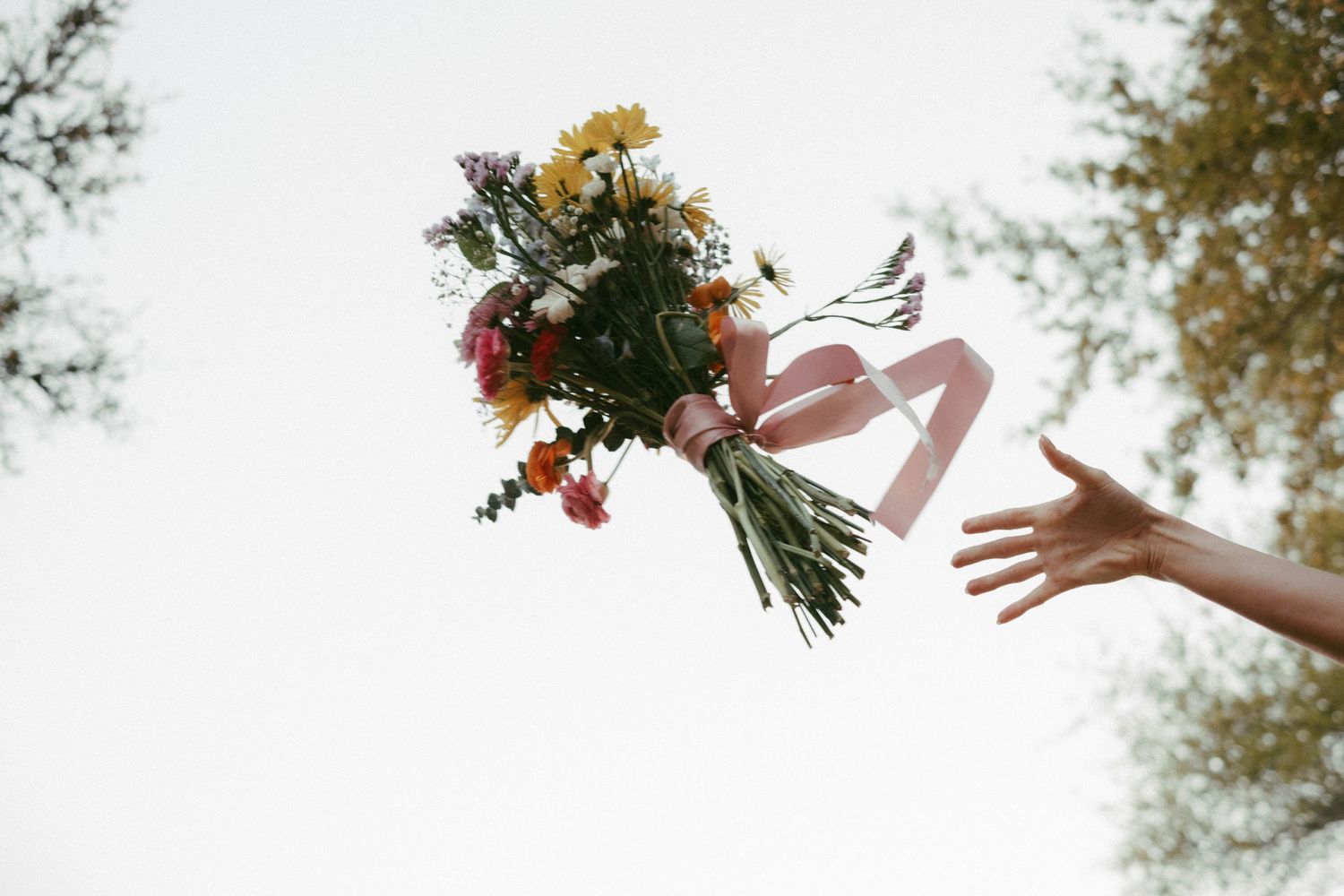 Wedding bouquet being tossed in the air with autumn trees and sunlight in the background.