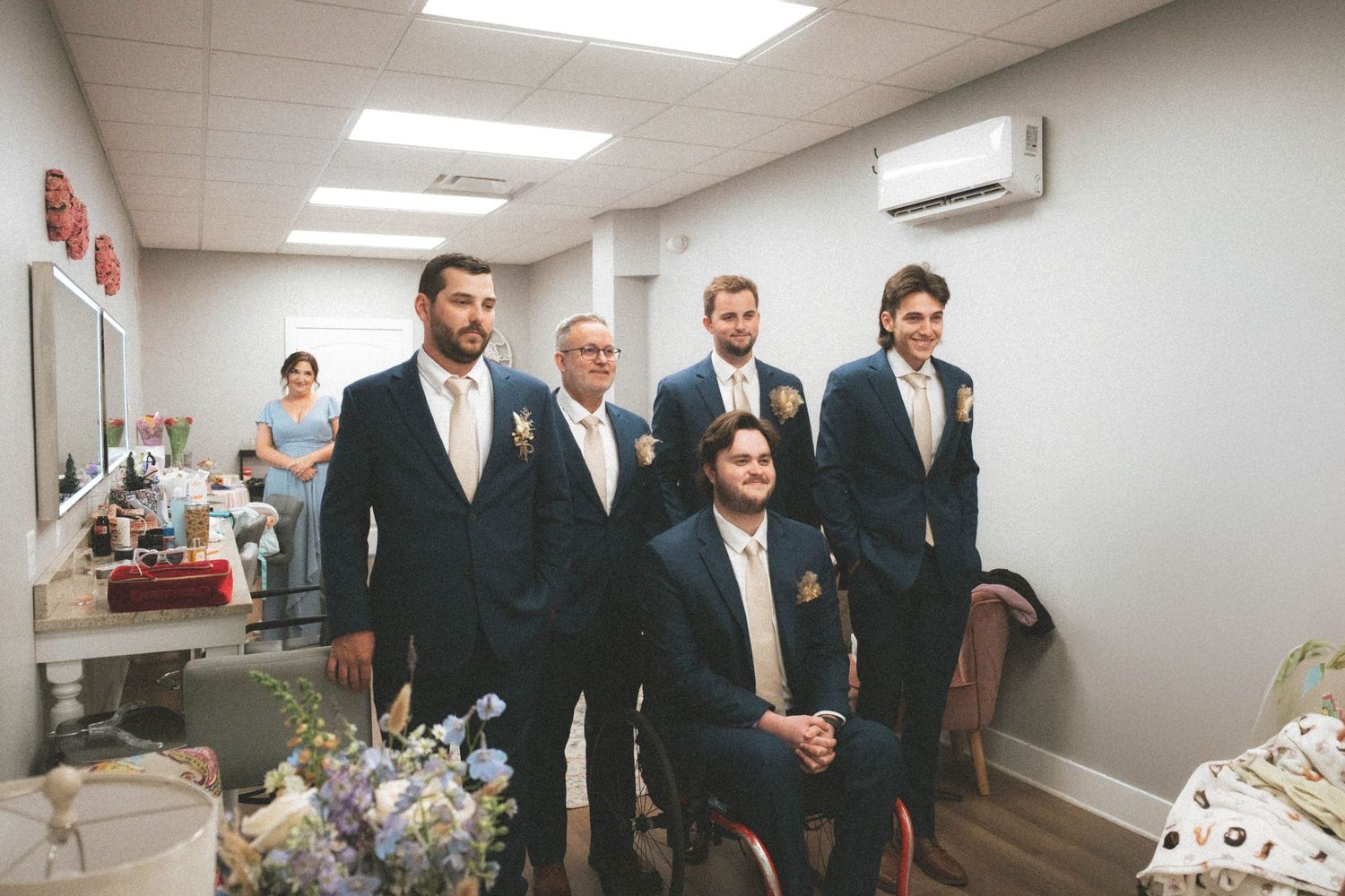 Five groomsmen in navy suits with boutonnieres pose together in a wedding preparation room.