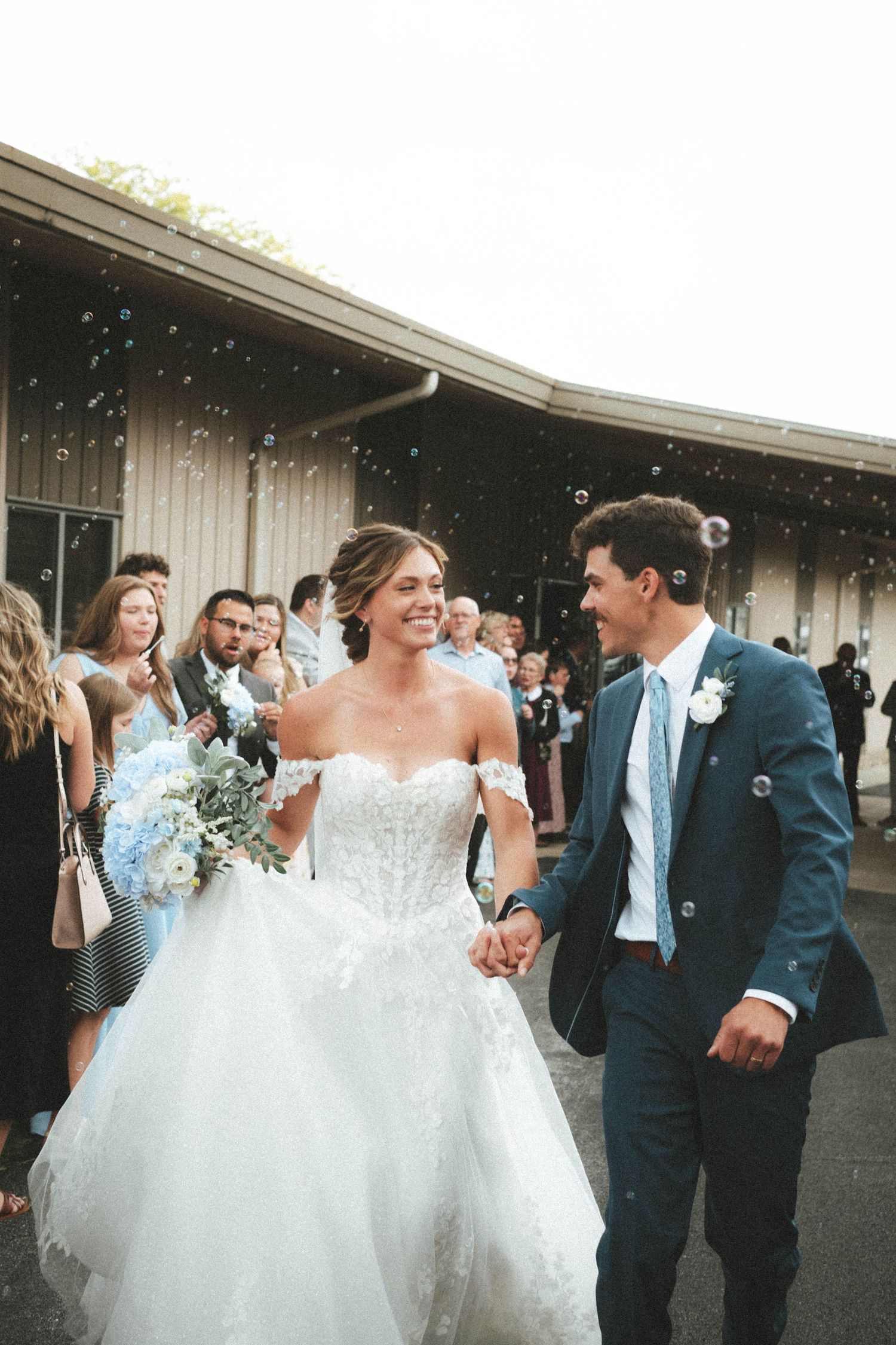 Smiling bride in off-shoulder lace gown and groom in navy suit exit wedding ceremony through bubble send-off.