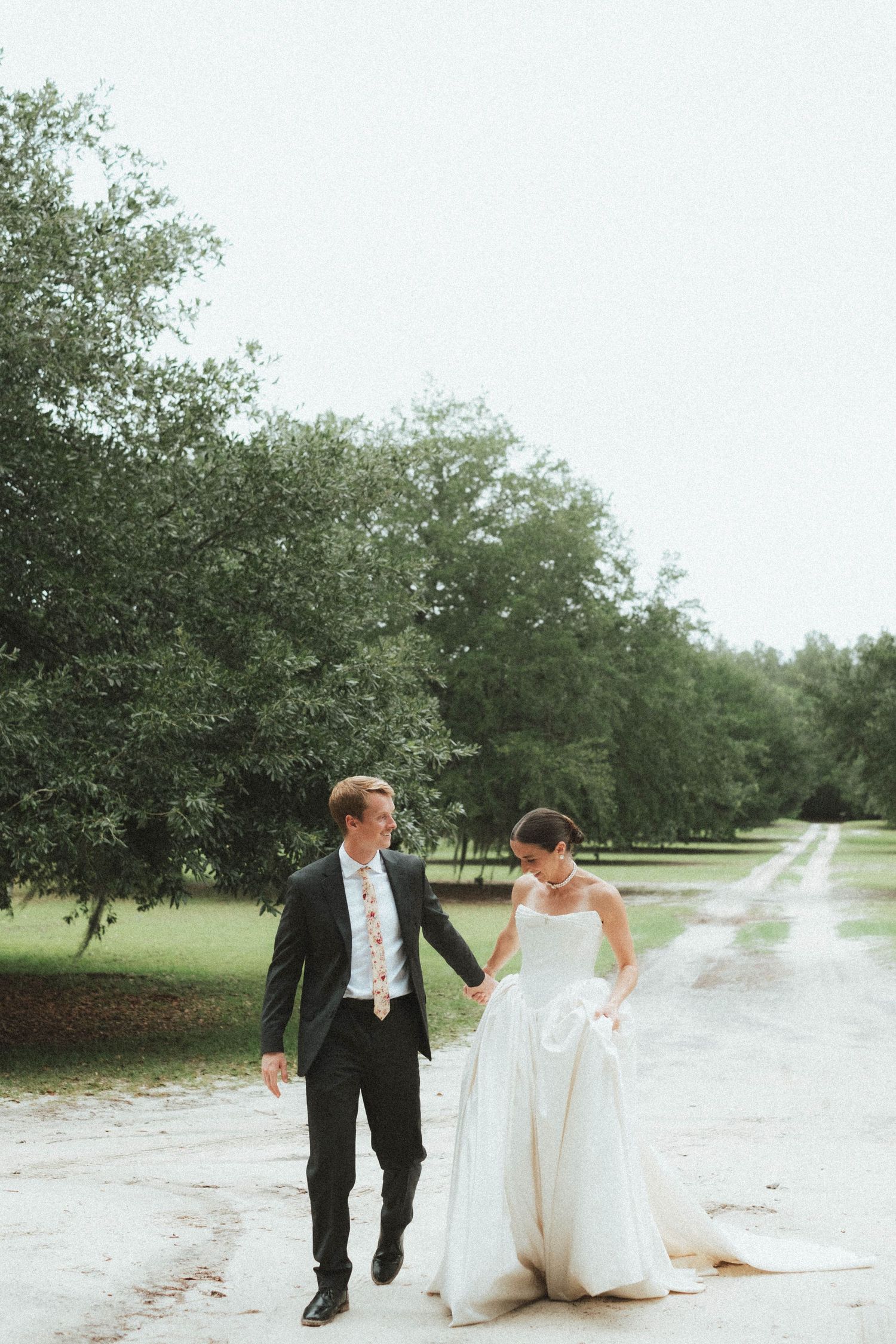 A bride and groom walk hand in hand down a tree-lined path, she in a white gown, he in a dark suit.