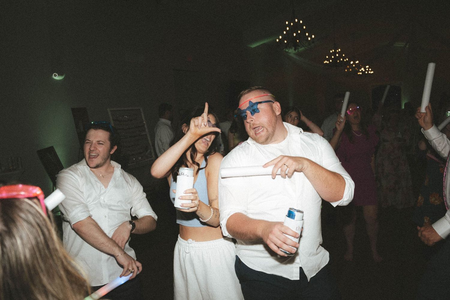 Guests dancing energetically at a wedding reception party, wearing white outfits and blue sunglasses under festive lights.