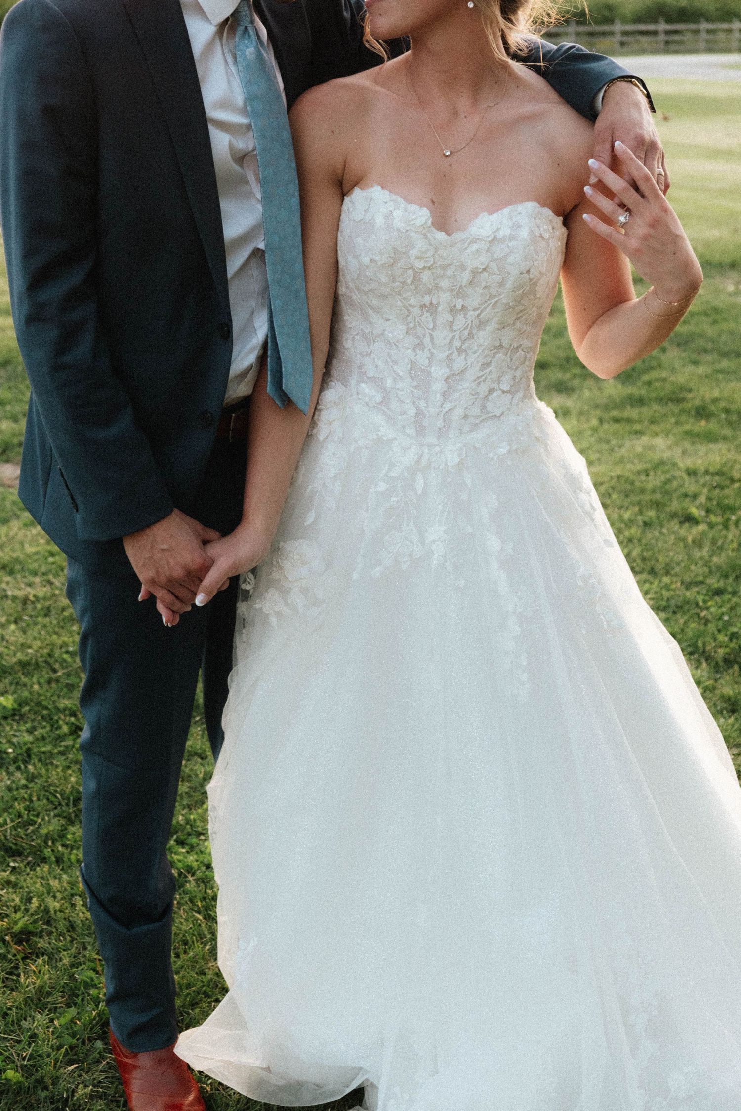 Close-up of bride in strapless lace corset wedding gown holding hands with groom in dark suit outdoors.