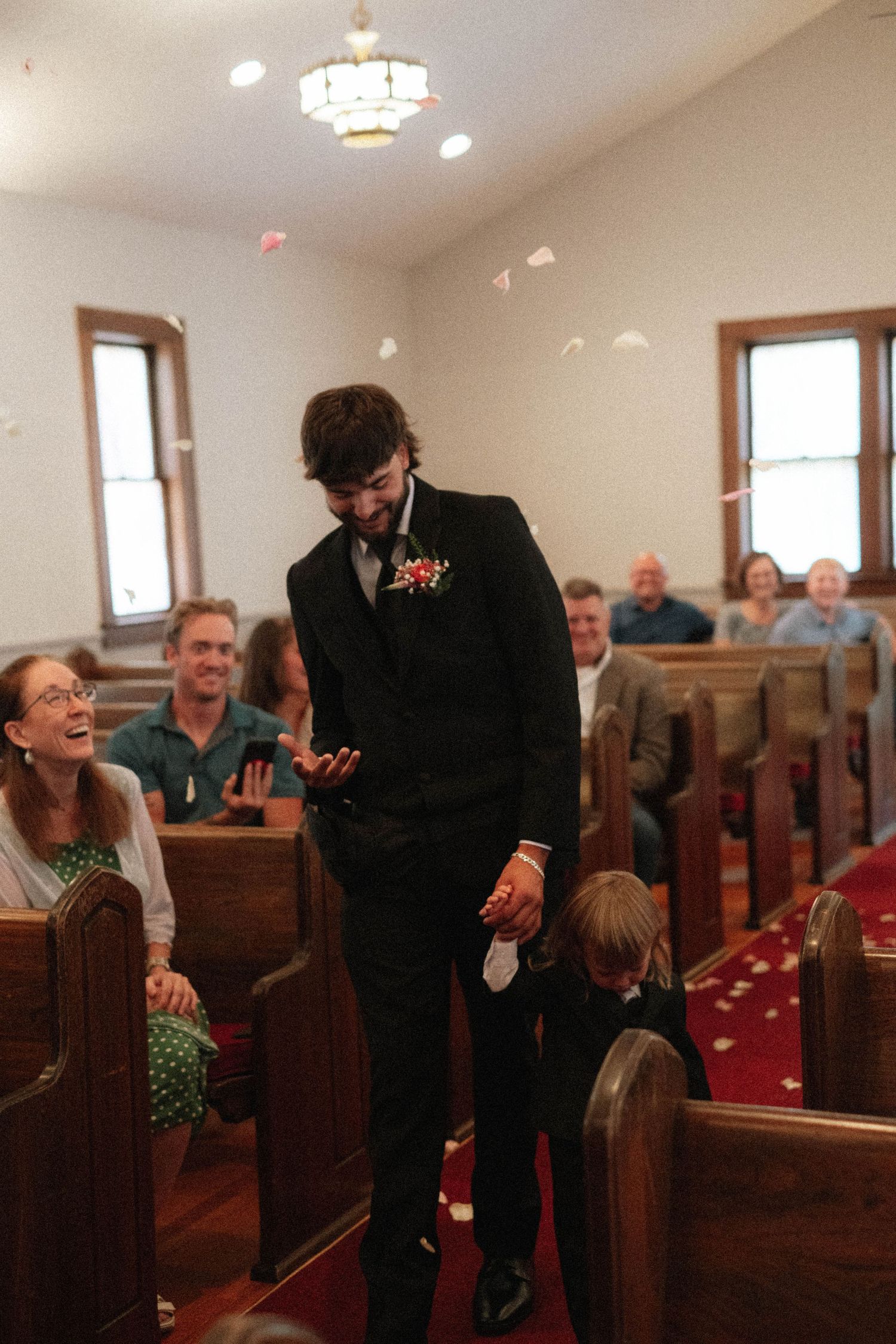 Groom in black suit walks down church aisle with young ring bearer as guests smile and flower petals fall.