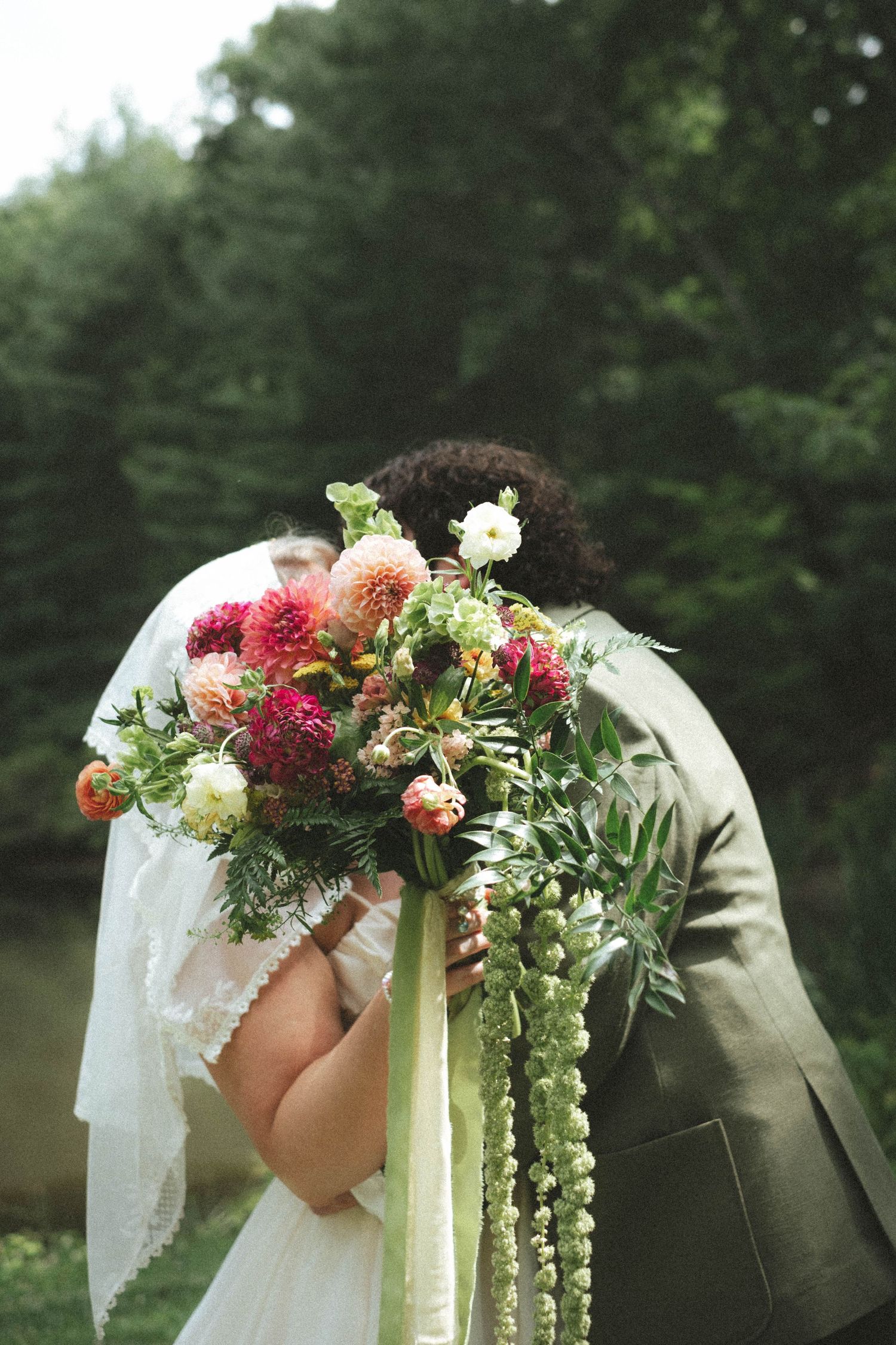 A bride and groom kissing behind a lush wedding bouquet of dahlias and greenery outdoors.