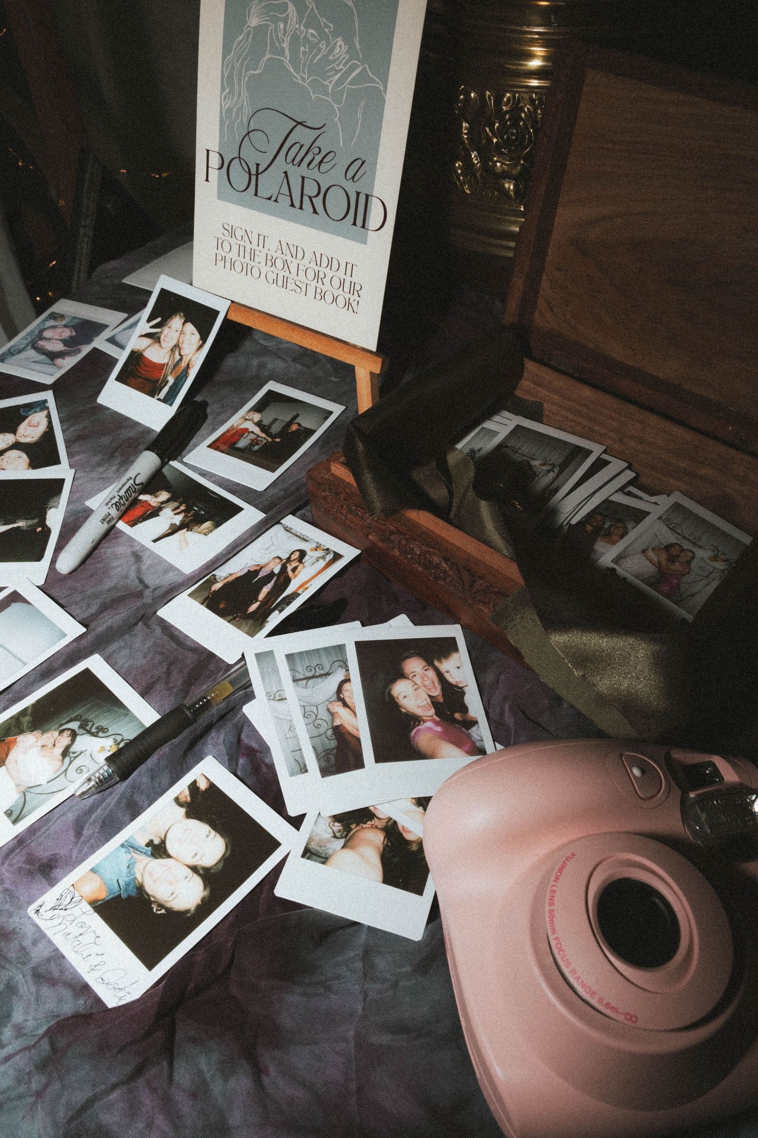 Pink Instax camera beside scattered Polaroid photos on a table with a Take a Polaroid guestbook sign at a wedding.