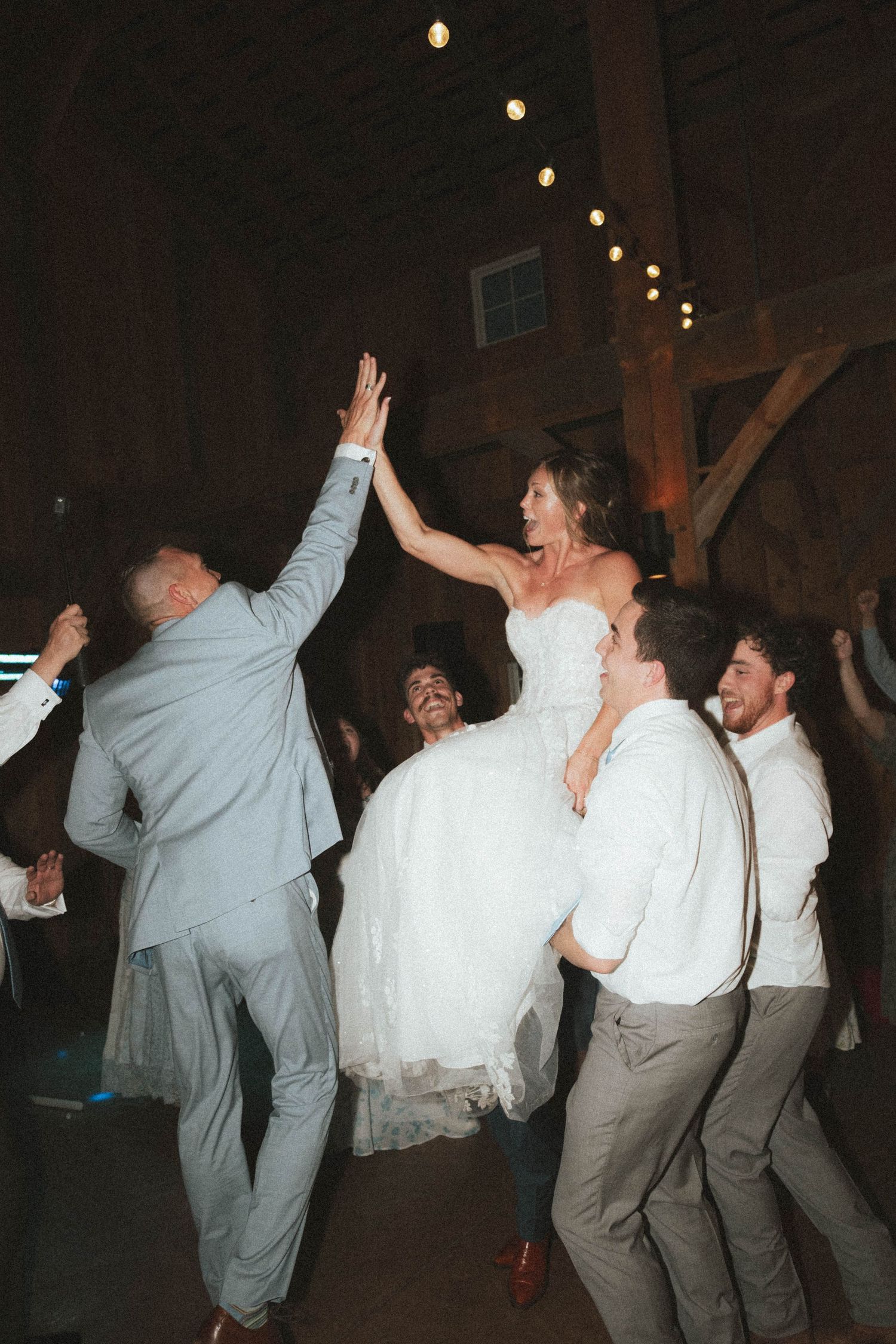 Bride in white gown lifted by groomsmen high-fiving during lively barn wedding reception celebration.