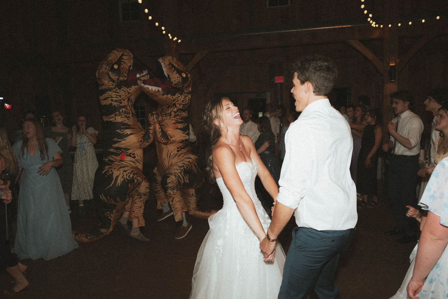 Bride and groom dance together at wedding reception as guest in T-Rex dinosaur costume joins the celebration.