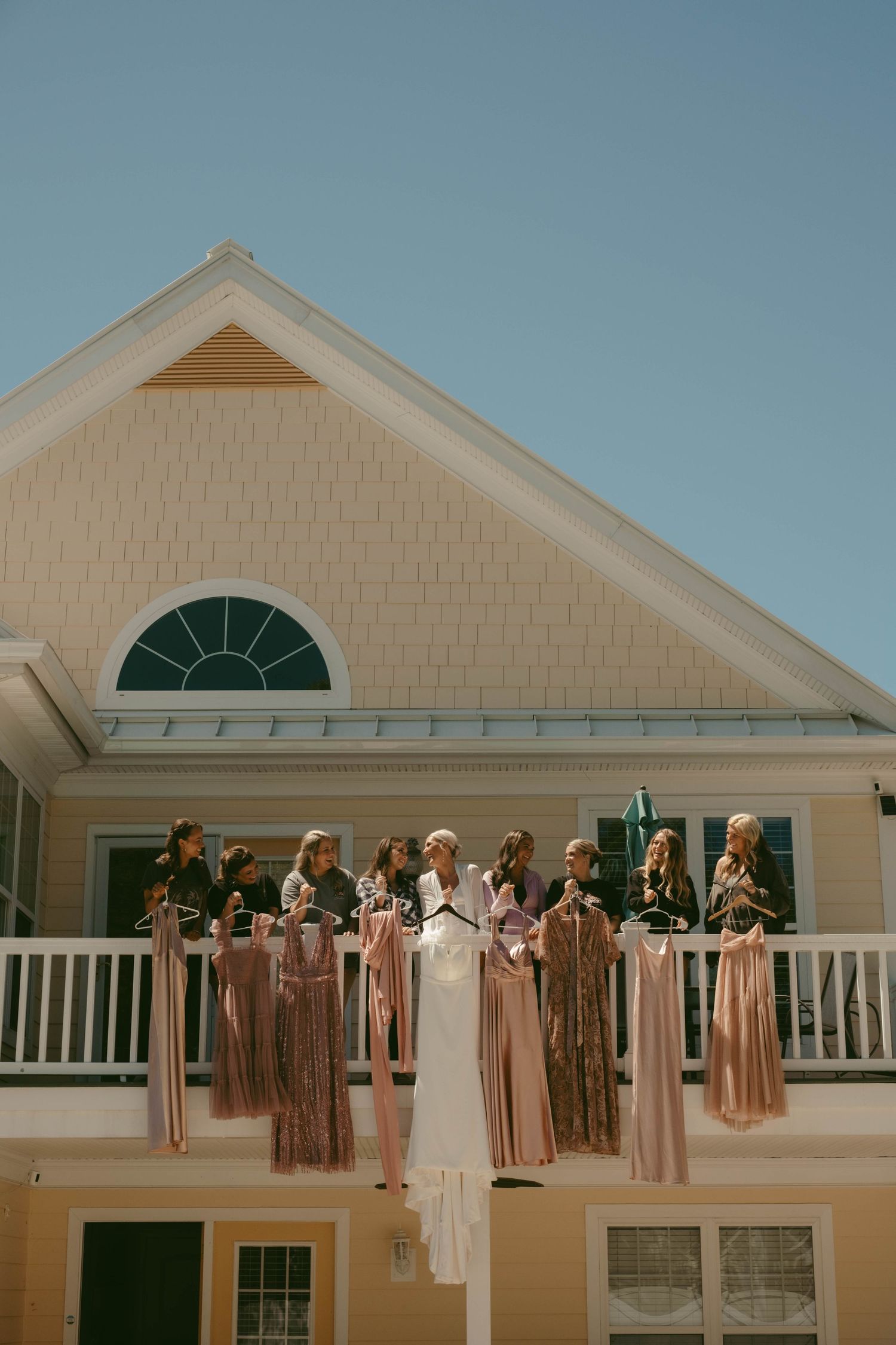 Row of blush and neutral colored bridesmaid dresses hanging outside a white house.