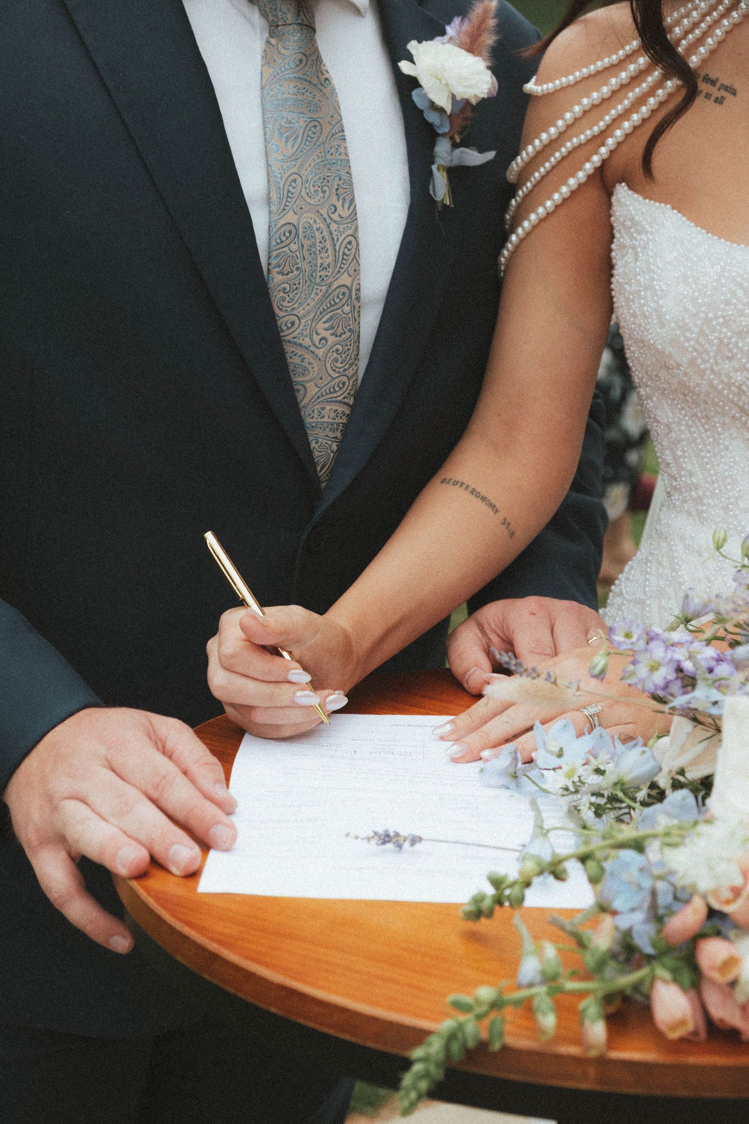 Bride signing wedding certificate at wooden table with gold pen, groom beside her, floral bouquet in foreground.