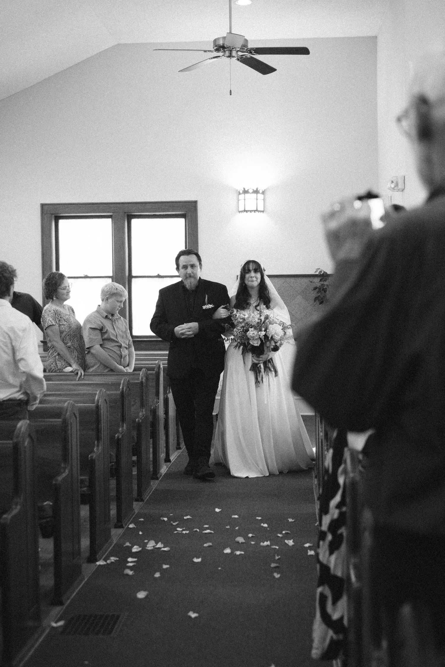 Black and white photo of a bride walking down the church aisle with her father during a wedding ceremony.