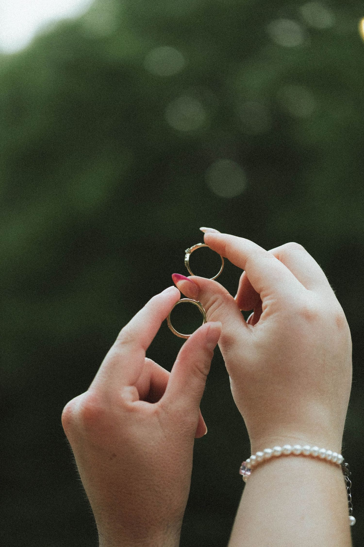 Two hands holding gold wedding rings outdoors, with pearl bracelet and pink nails visible against green bokeh background.