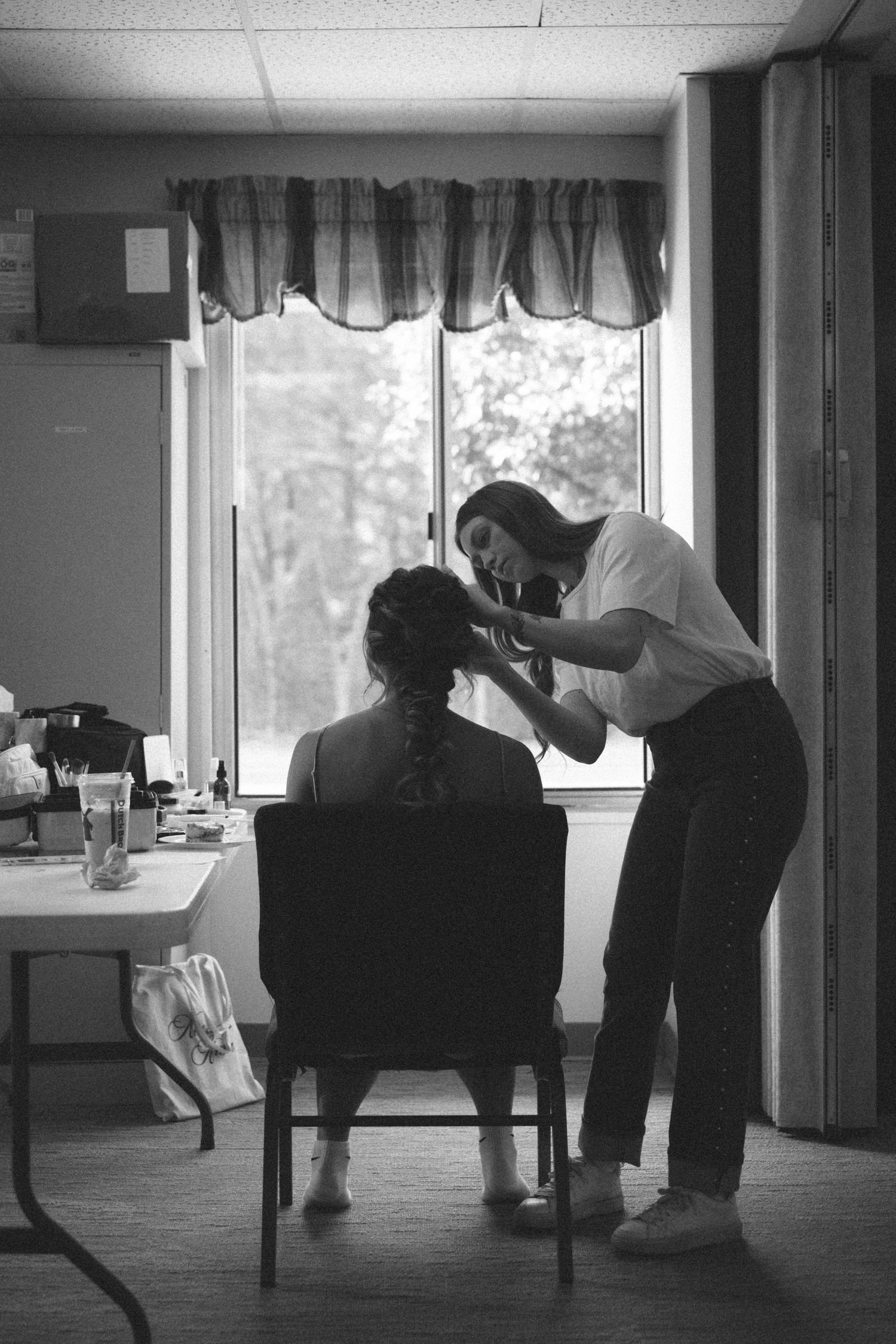 Black and white photo of a makeup artist applying makeup to a seated woman near a window indoors.