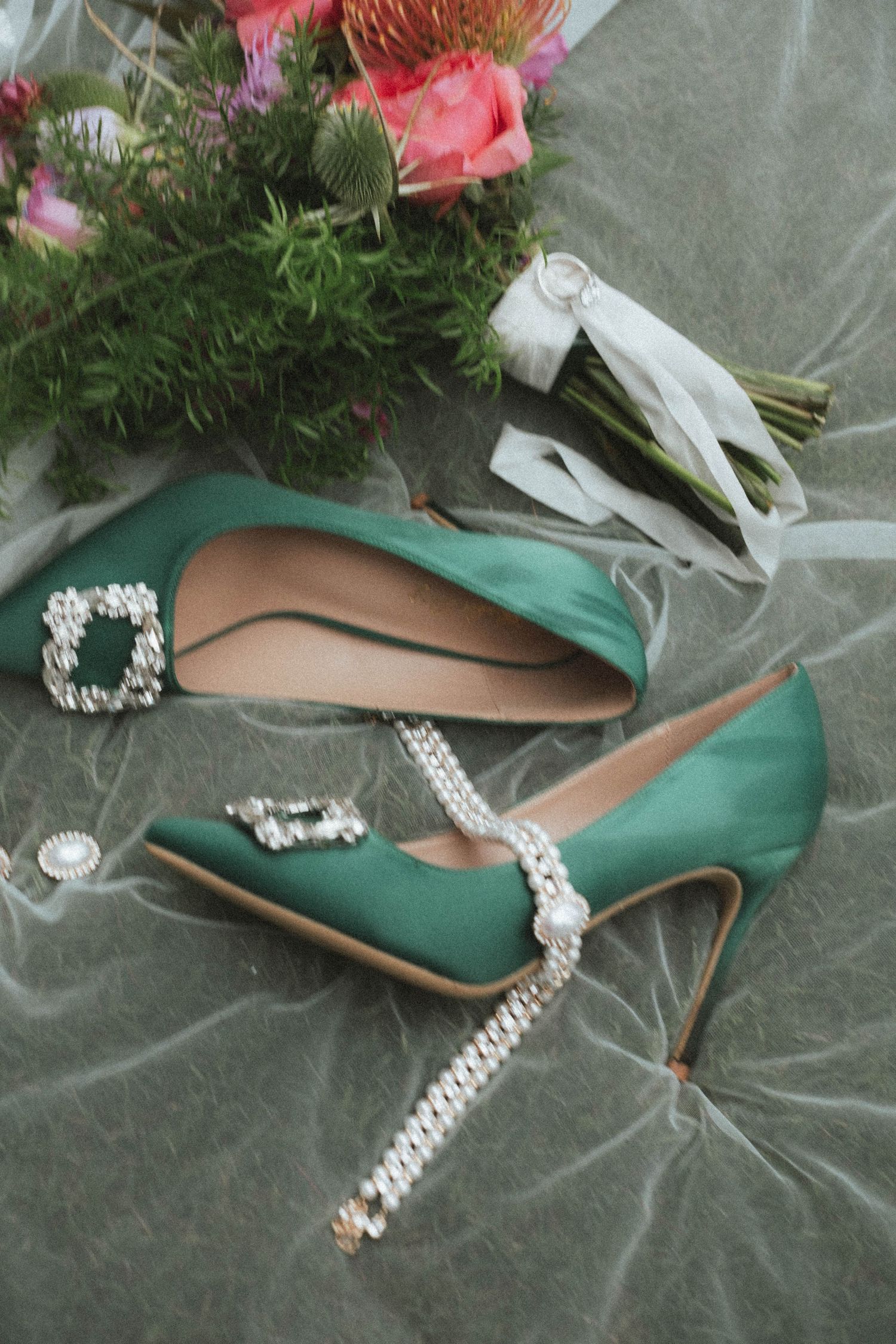 Elegant green satin stiletto heels with crystal buckle embellishments displayed beside a floral bouquet on gray velvet.