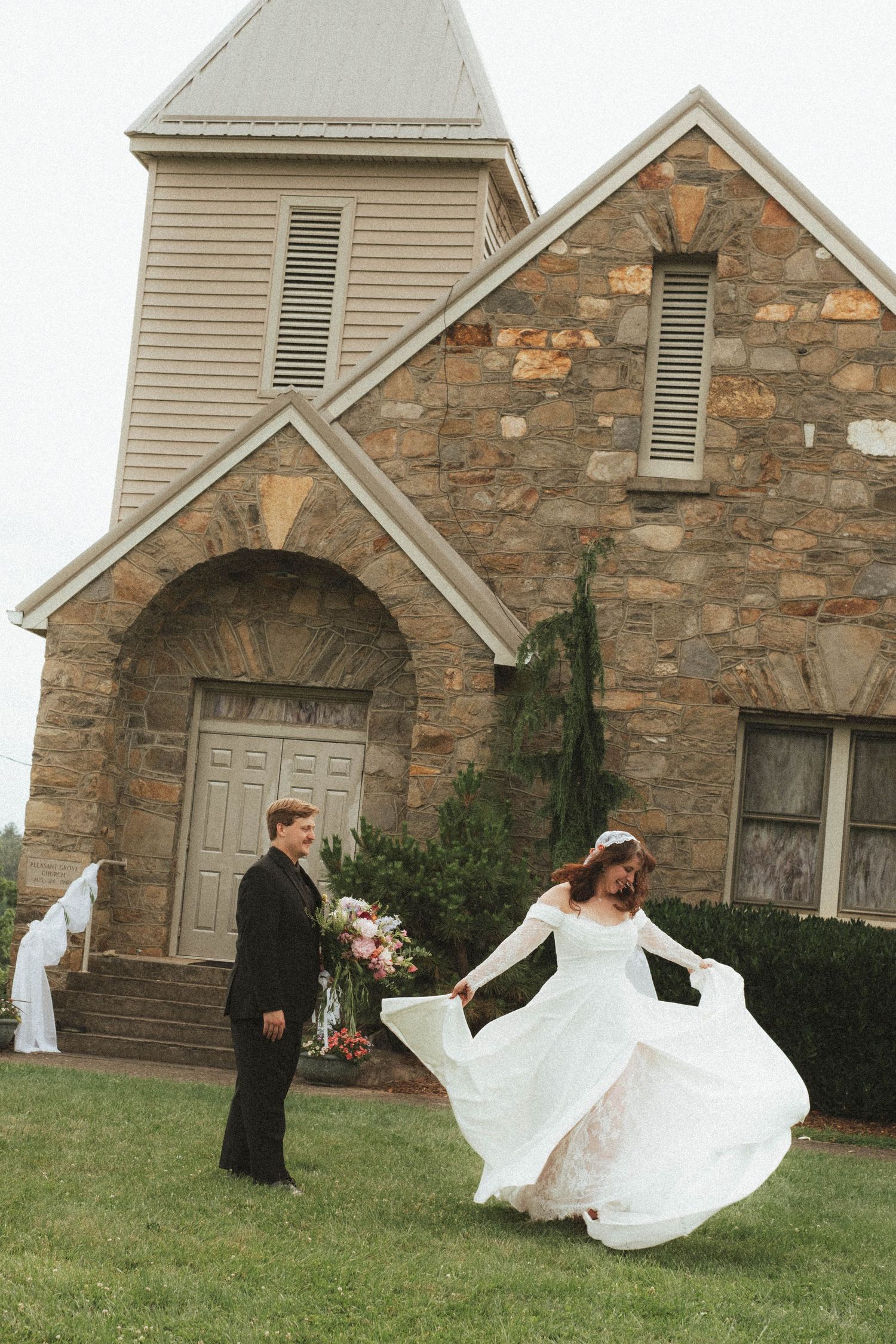 Bride twirling in white gown outside stone church with groom in black suit holding bouquet on wedding day.