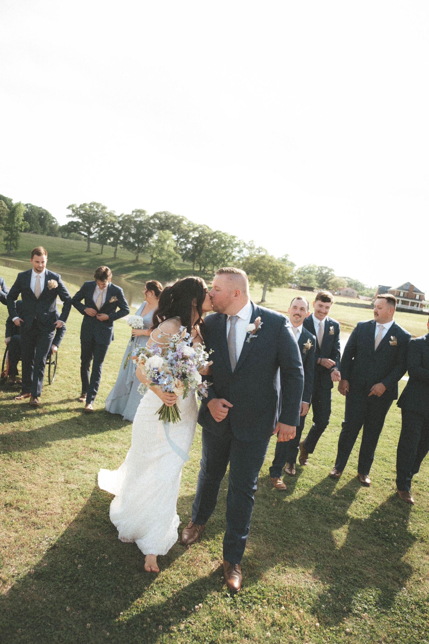 Bride and groom share a kiss while wedding party walks behind them on a sunny green lawn.