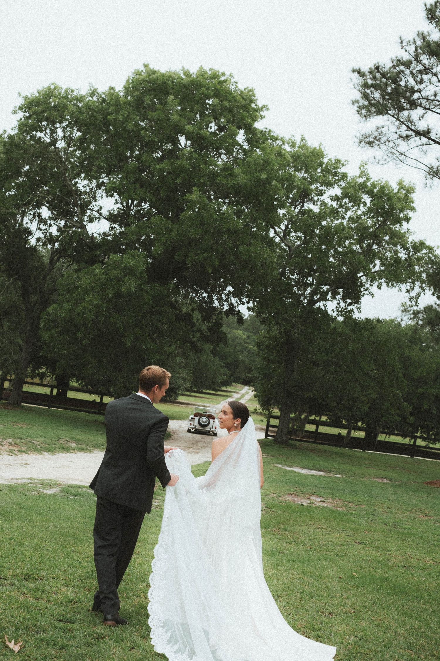 Bride and groom walk hand-in-hand through lush green grounds, her white gown and veil trailing behind them.