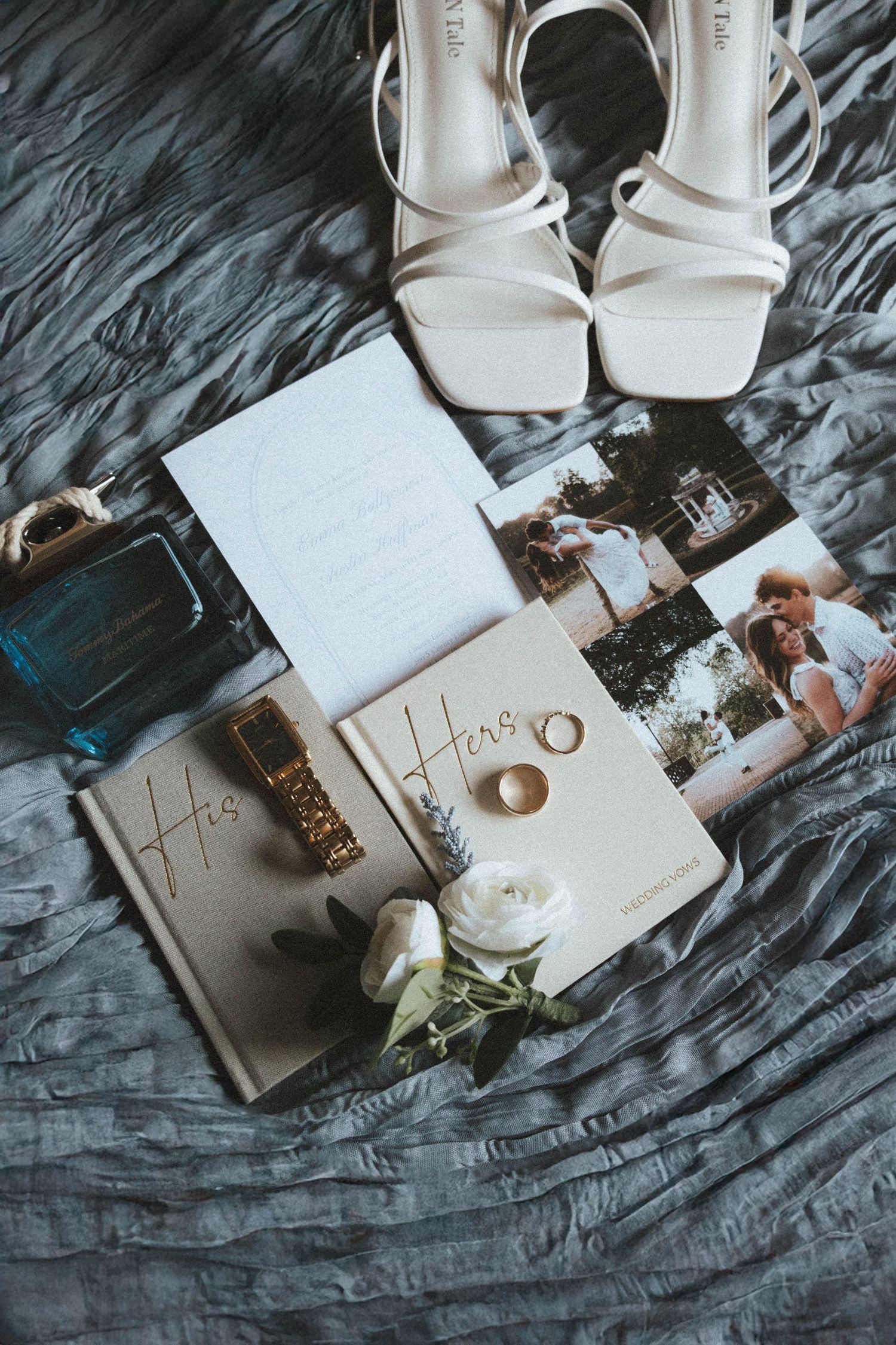 Elegant wedding flat lay with white heels, gold rings, His and Hers books, perfume, watch, and floral invitation on gray fabric.