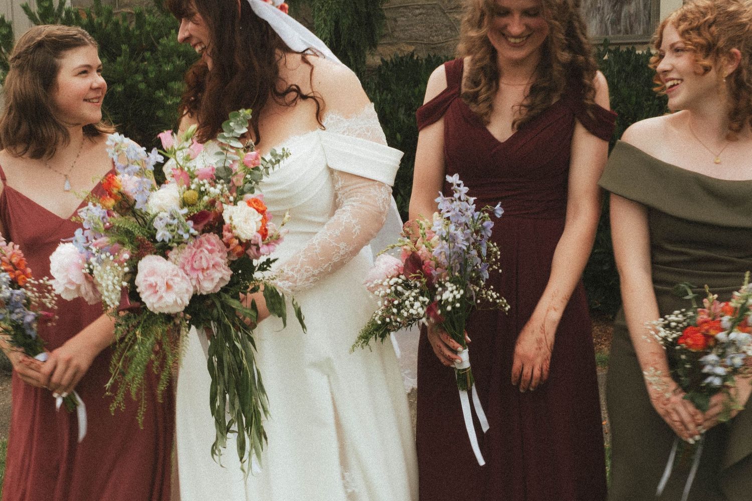 Bride in white gown with bridesmaids in mismatched burgundy and sage dresses holding colorful wildflower bouquets outdoors.