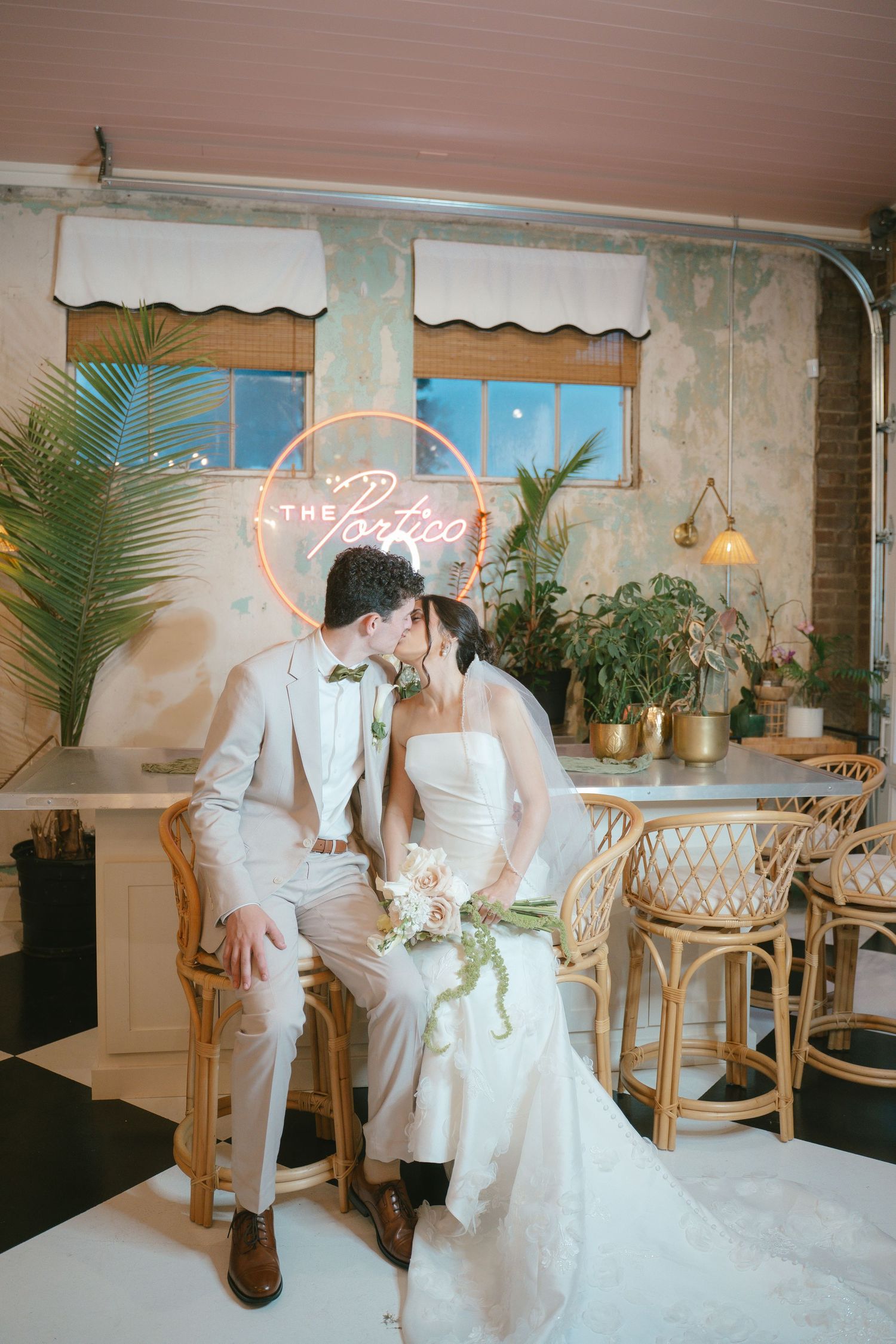 Wedding couple in white attire seated in a vintage-inspired room with rattan furniture and tropical decor.