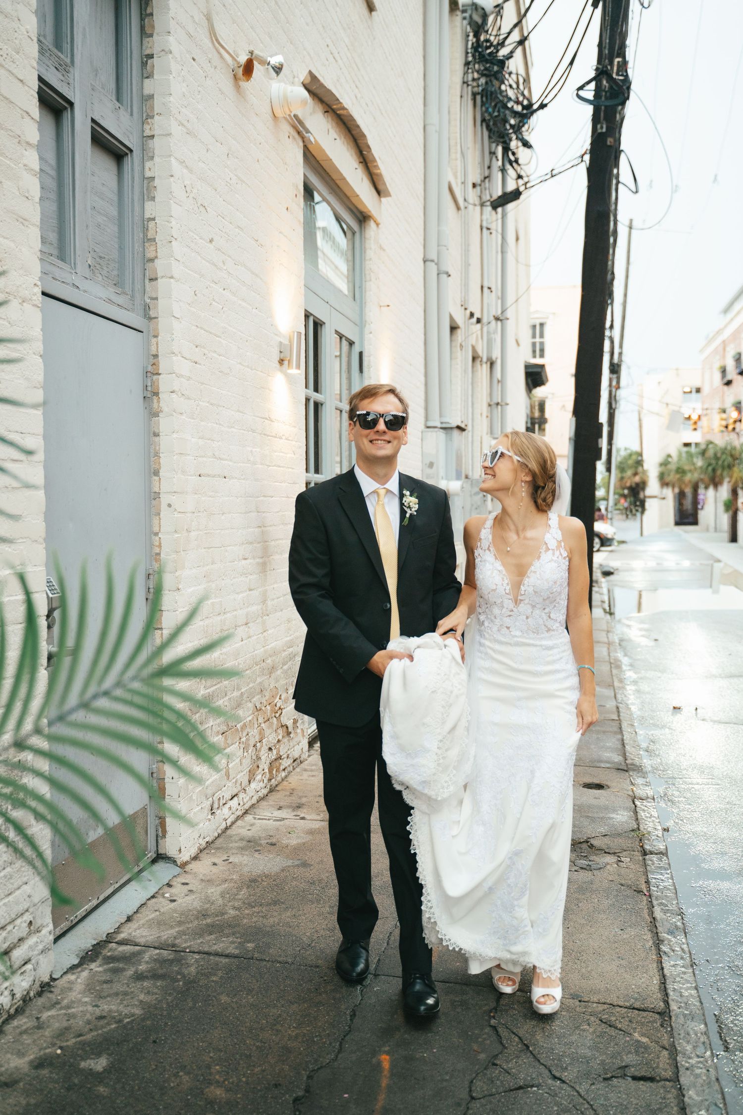 Bride and groom in sunglasses walking down a charming urban alley, she in lace gown, he in black suit with yellow tie.