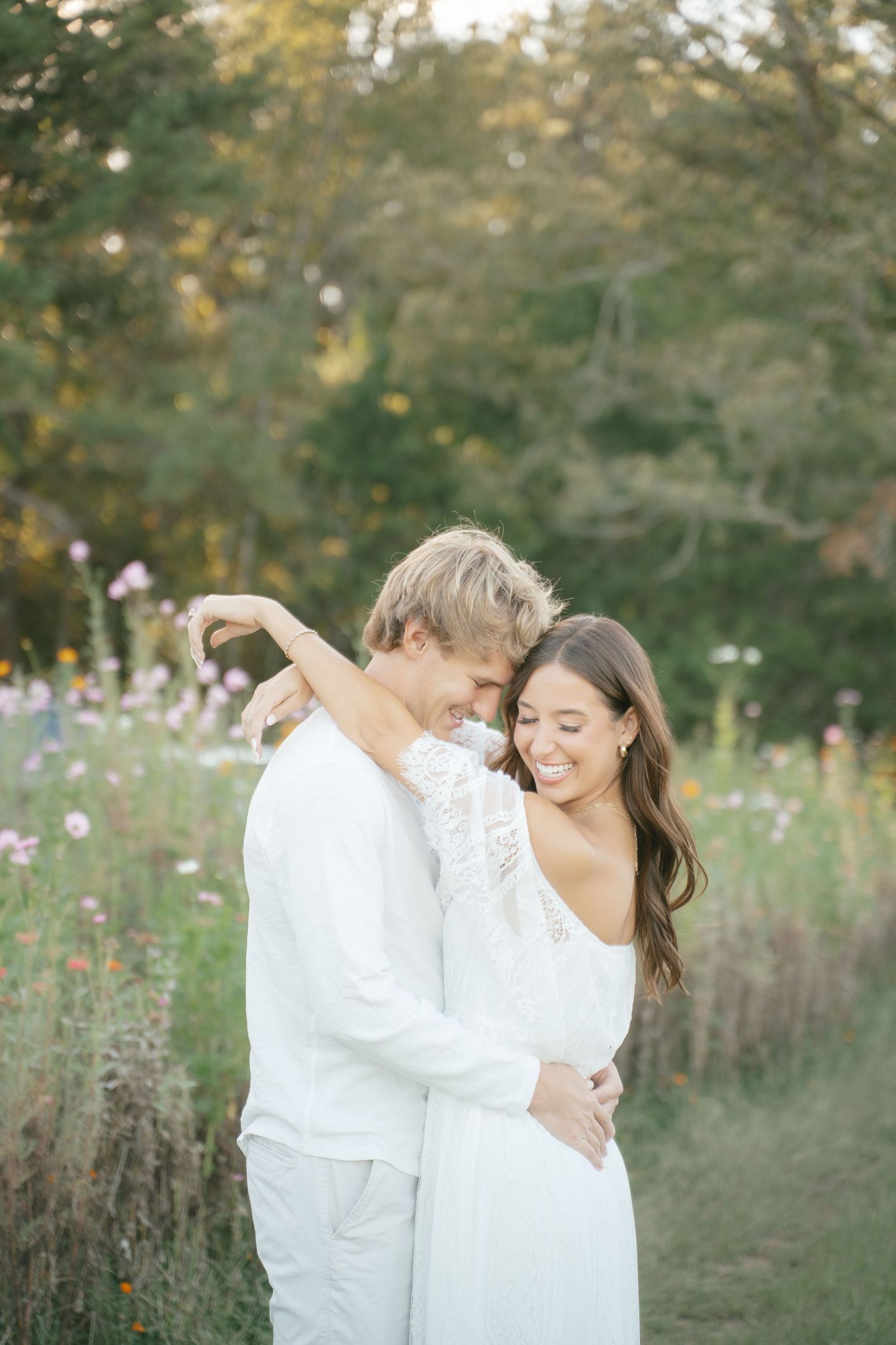 A couple in white outfits embraces in a wildflower field during golden hour.