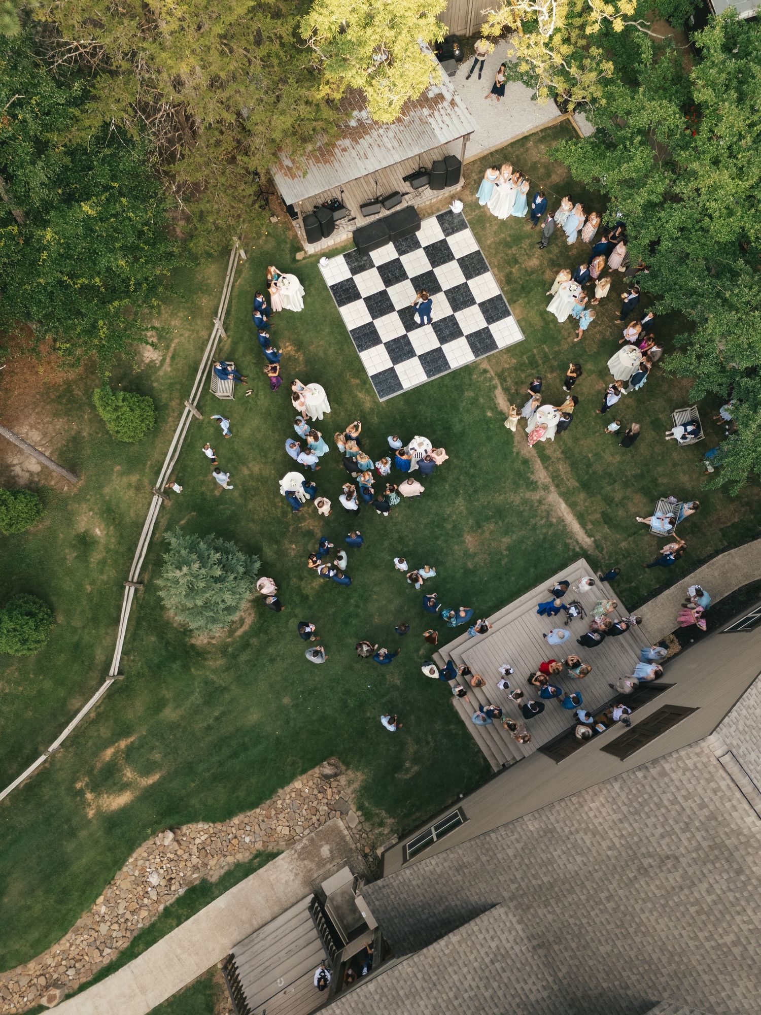 Aerial view of a backyard gathering with a giant chess board on green lawn surrounded by trees.
