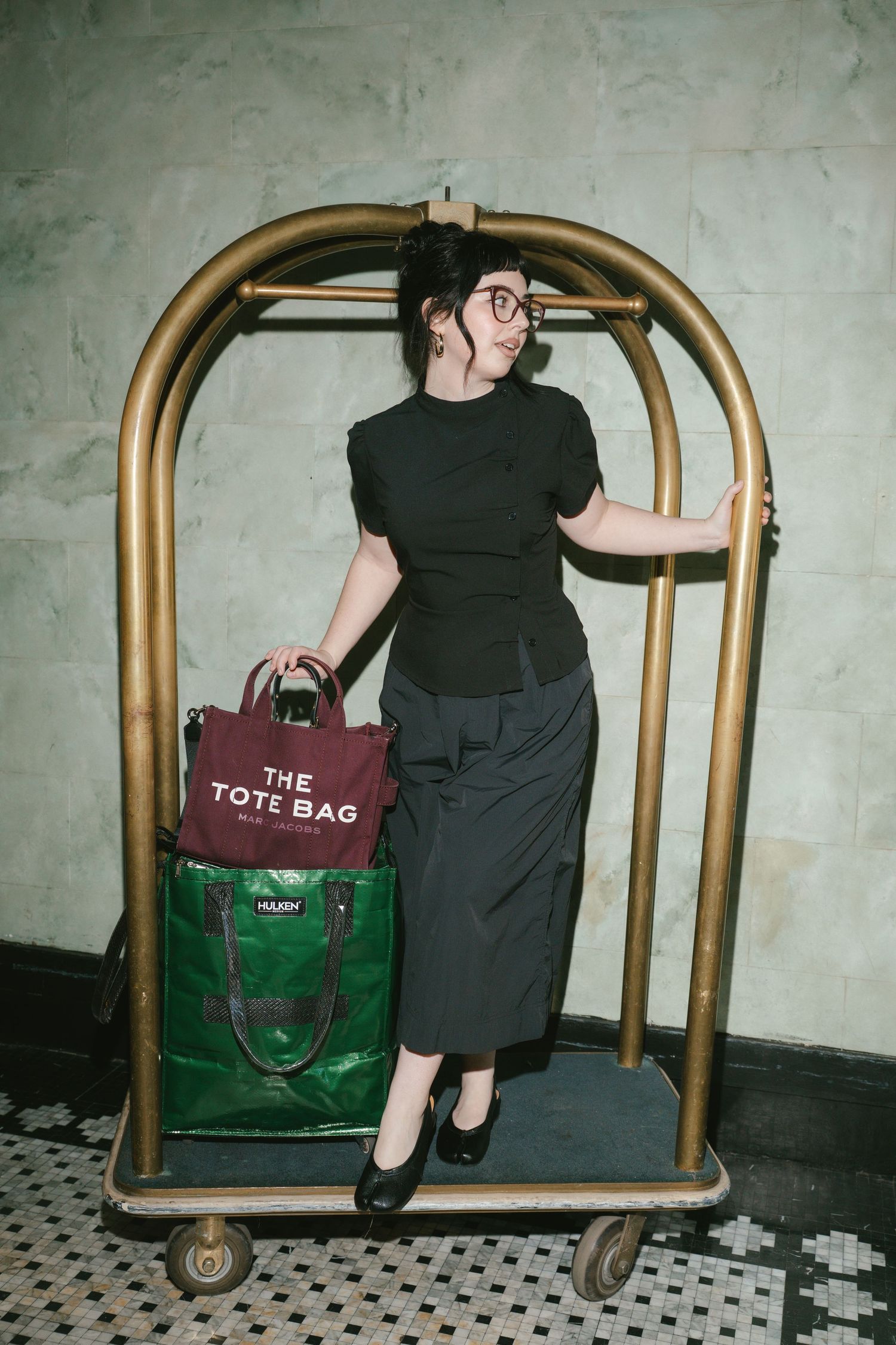 Woman in black outfit holding Marc Jacobs tote bag, posing on a gold hotel luggage cart indoors.