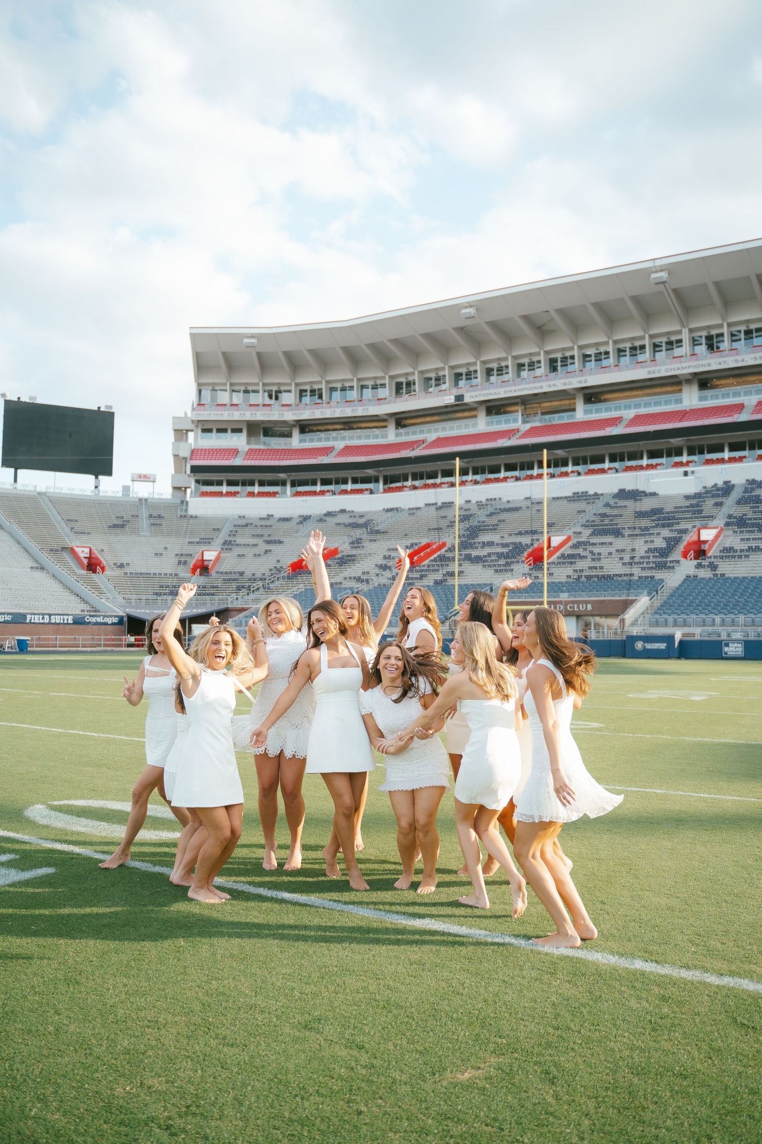 People in white outfits jump and pose together on a sunny day at a football stadium.